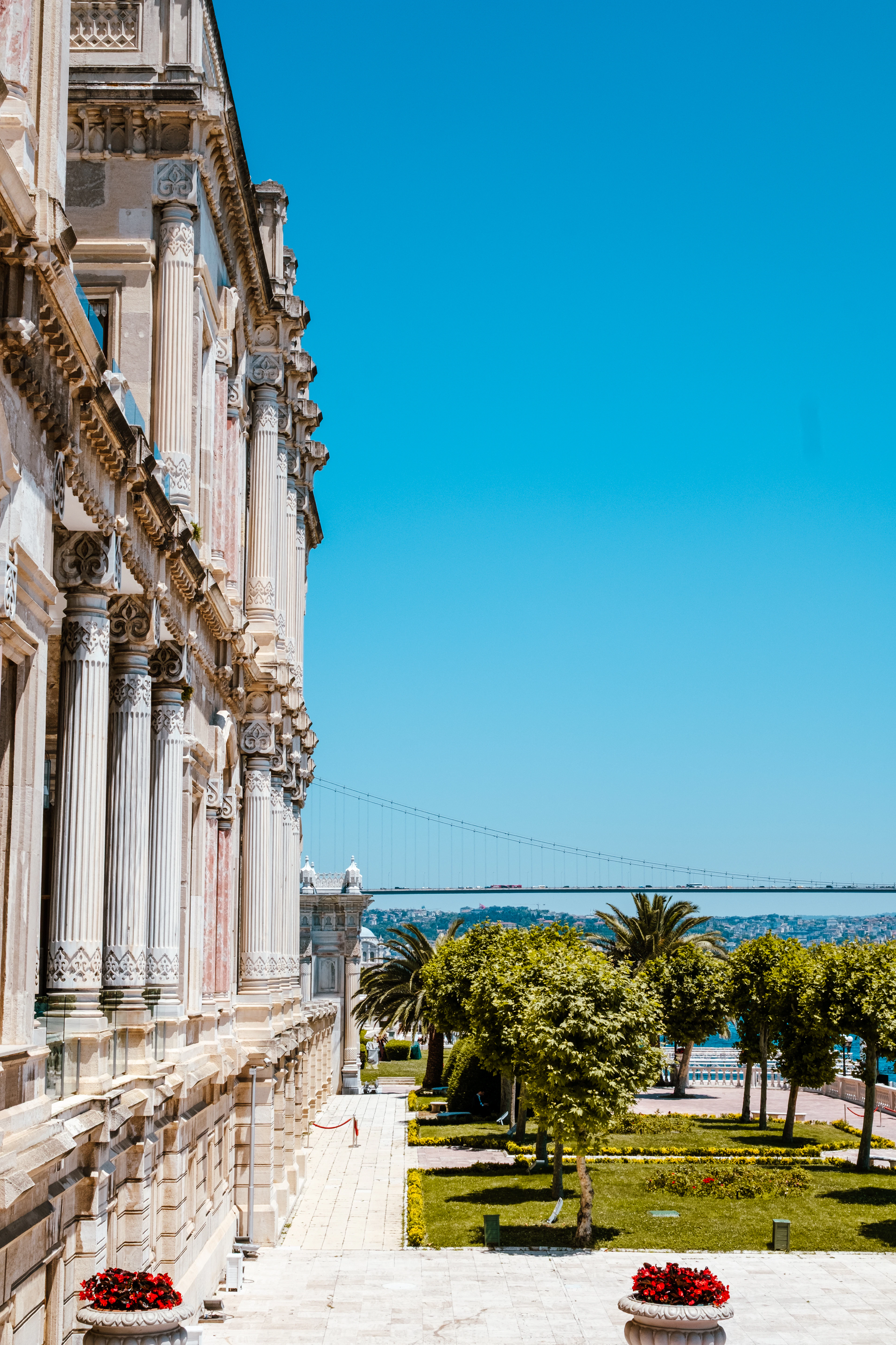 The carved marble facade of the Ciragan Palace on the Bosphorus in Istanbul seen along its terrace, palms and flowerbeds in front and the Bosphorus Bridge spanning the strait beyond