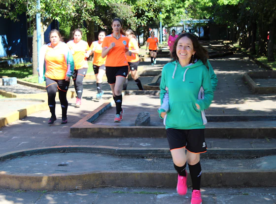 Grupo de mulheres correndo em treino de assessoria em parque arborizado, com camisetas laranja de equipe e paces diferentes