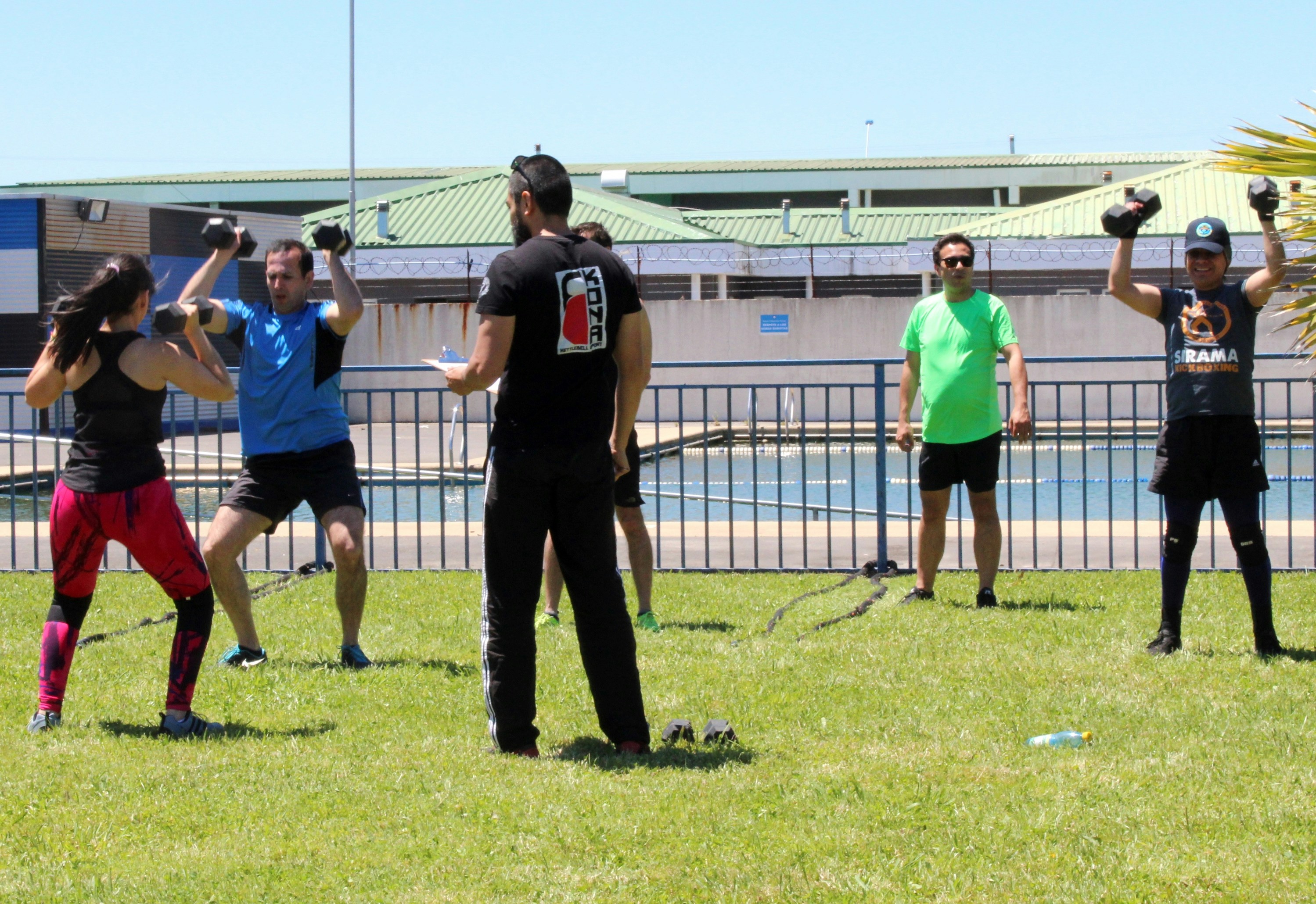 A group of people exercising with dumbbells on a grassy field near a pool
