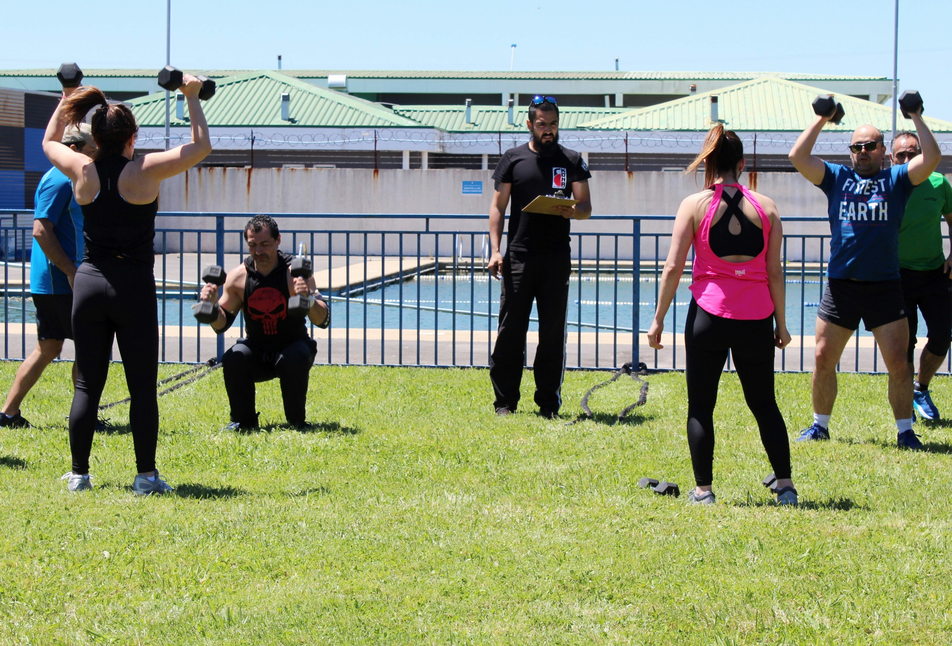A group of people exercising with dumbbells on a grassy field near a pool