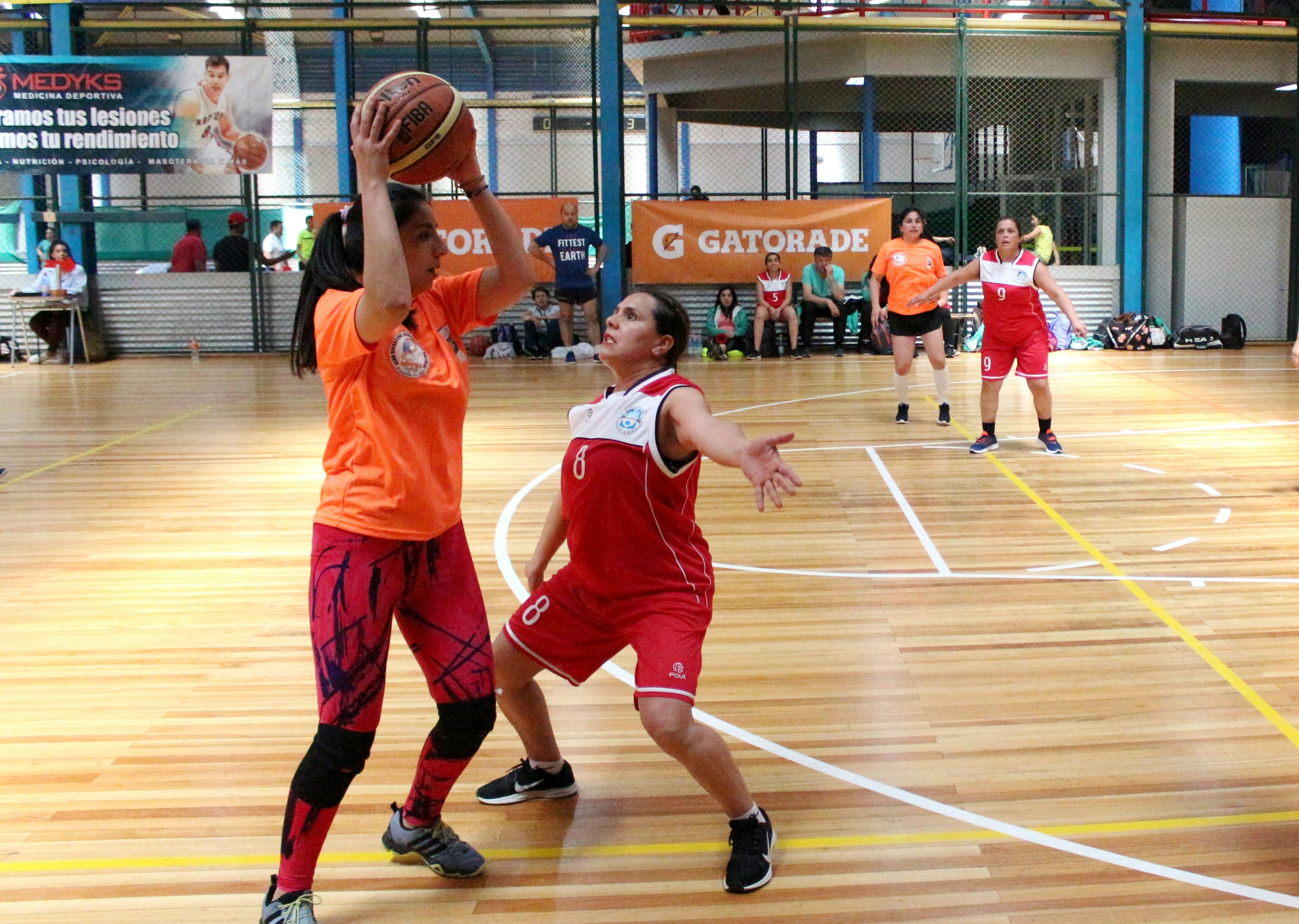 A woman in an orange shirt holding a basketball above a defending player