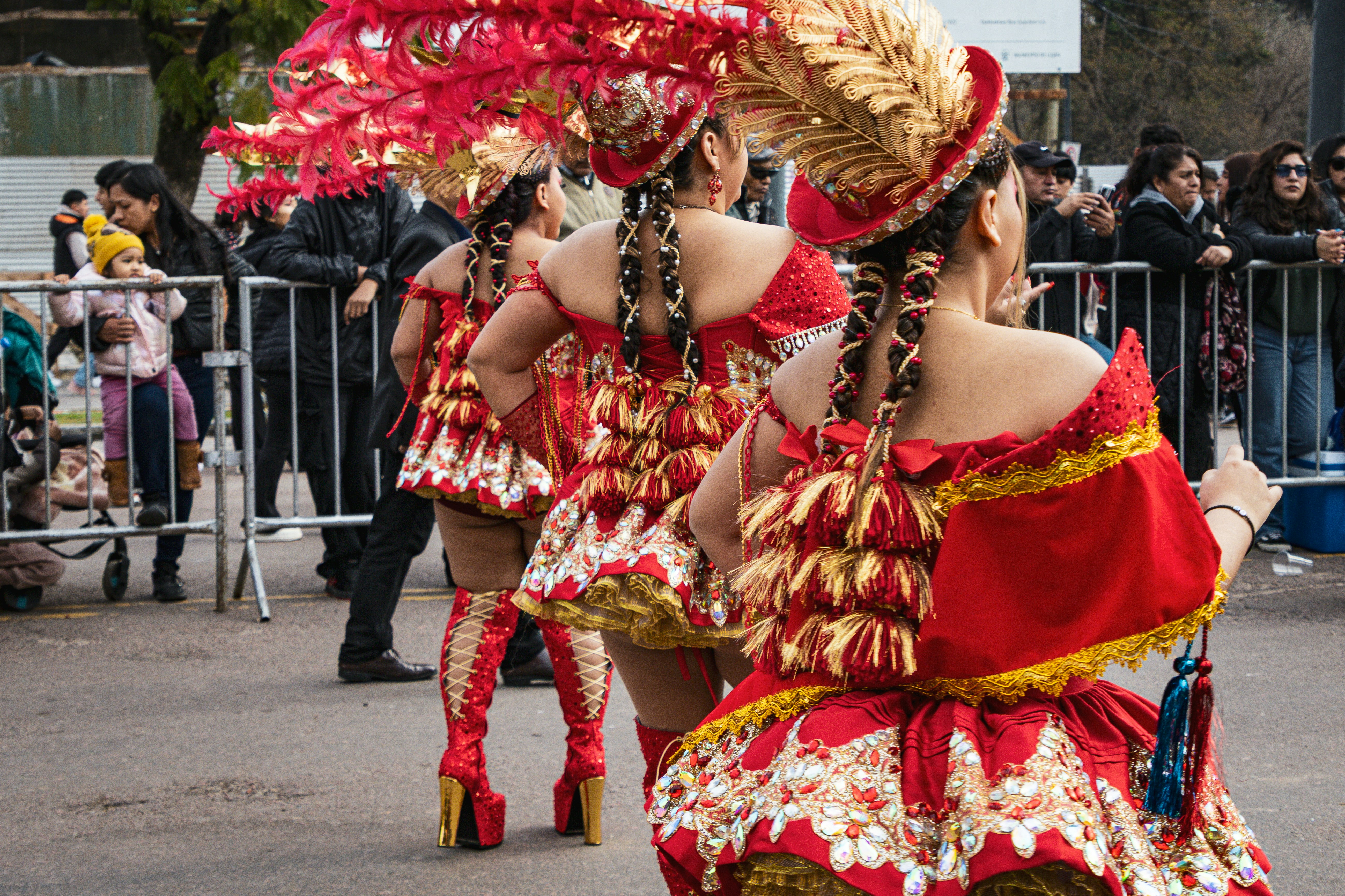 Três dançarinos em trajes ornamentados vermelhos e dourados com cocares de penas em um desfile.