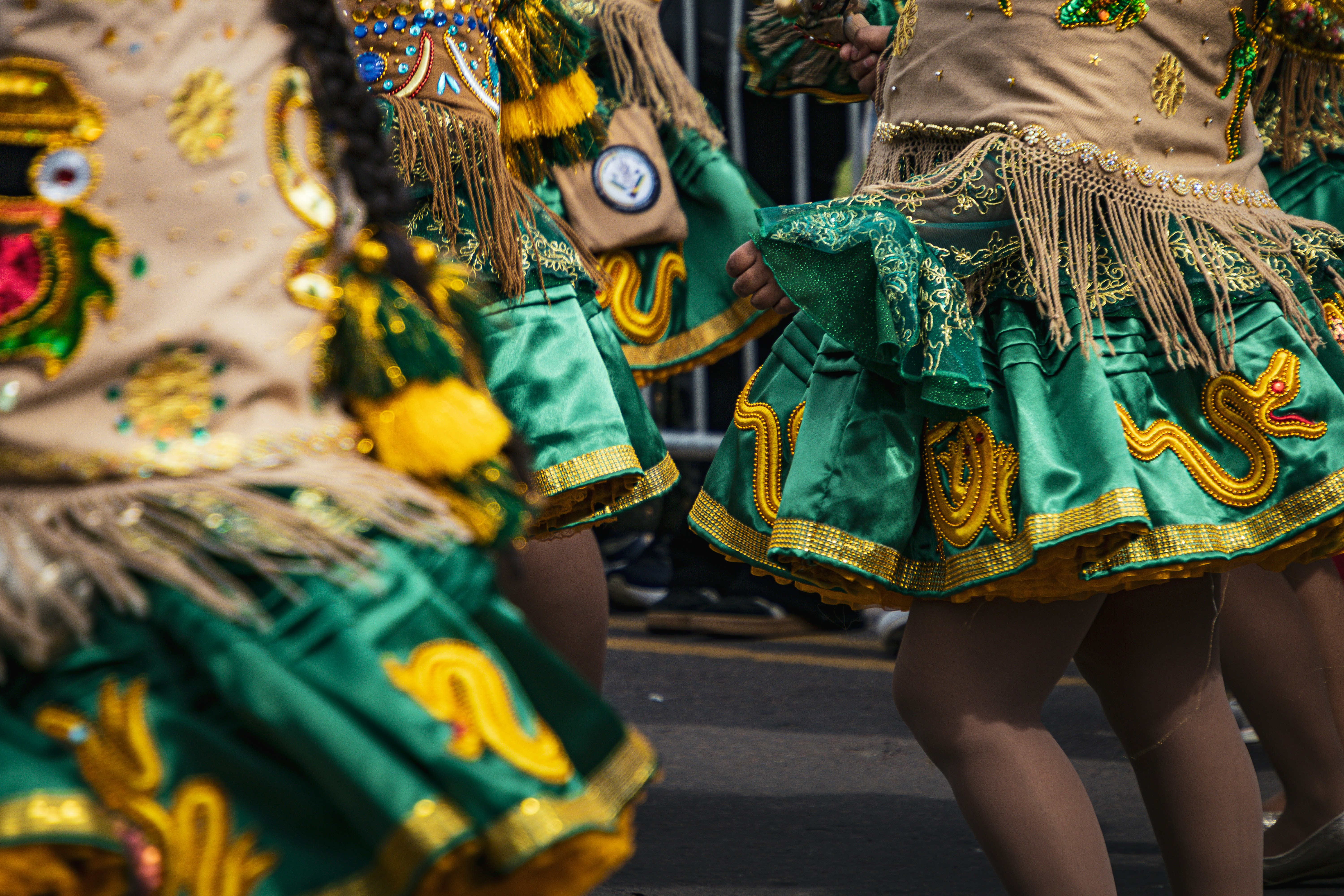 Dançarinos com saias de cetim verde e dourado com franjas se apresentando em um desfile de rua.