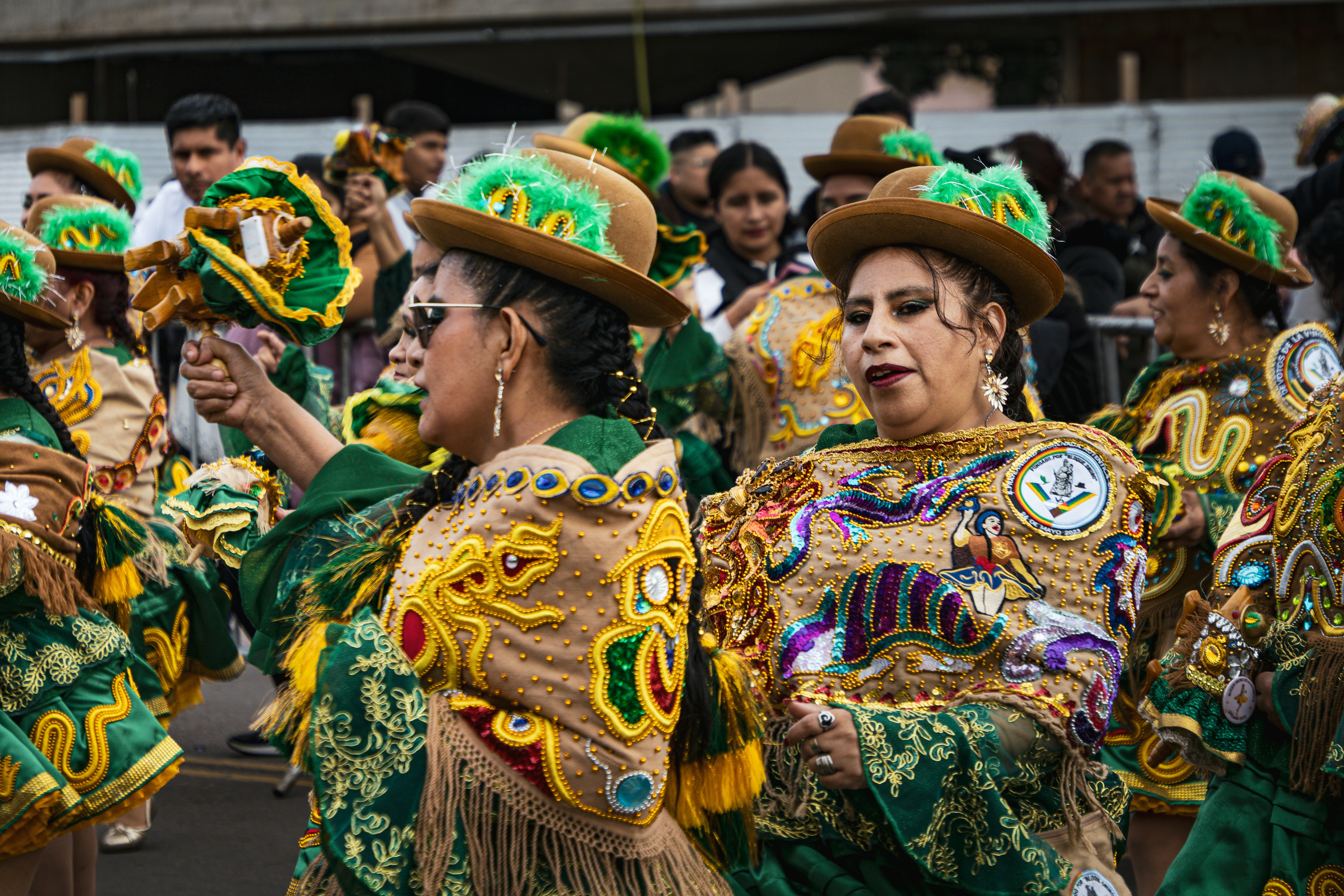 Dançarinos em trajes ornamentados verdes e dourados com chapéus se apresentando em um desfile de rua.