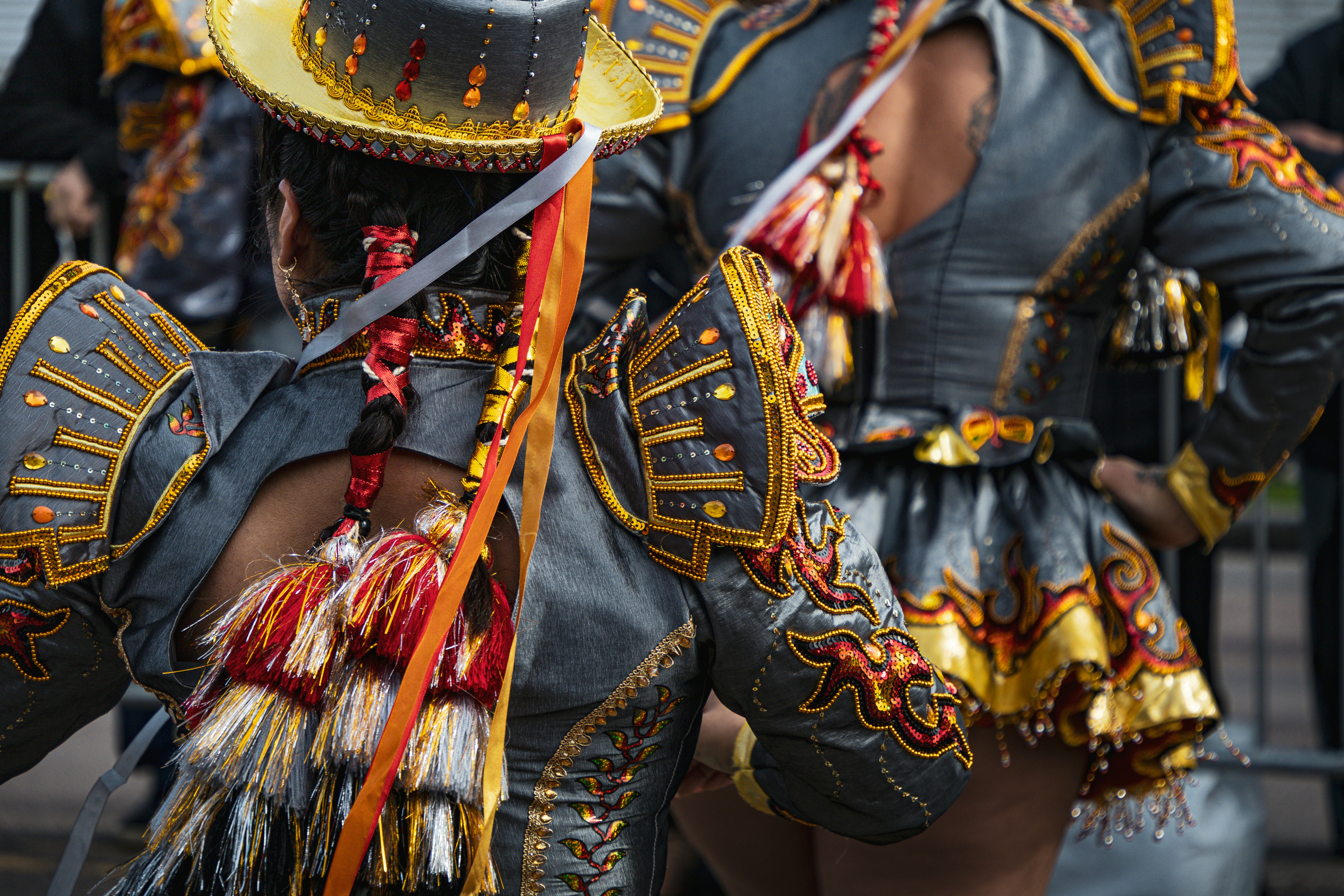 Duas pessoas em trajes ornamentados cinza e dourado com franjas em um desfile.