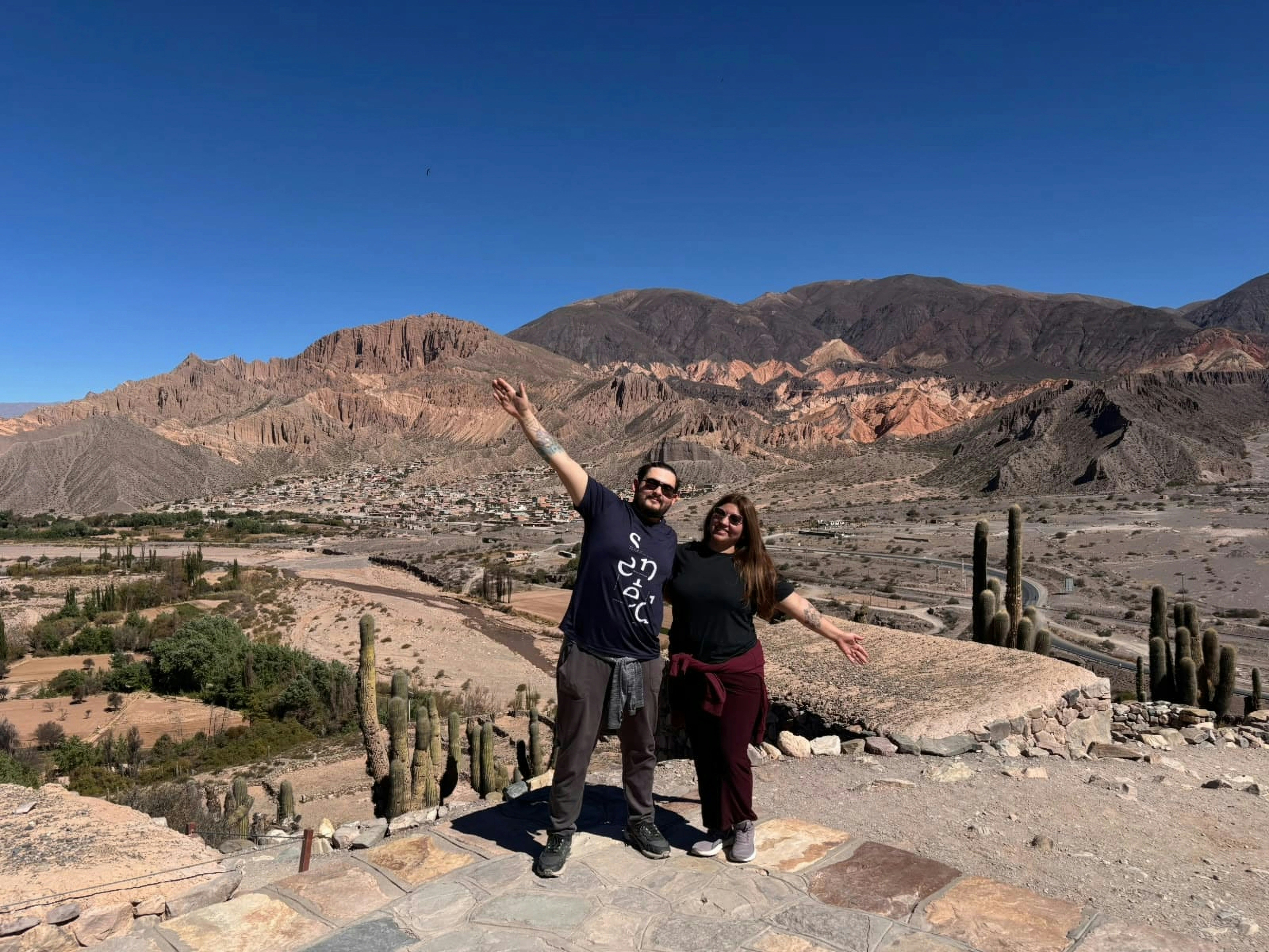 Two adults with arms outstretched on a rocky overlook above a desert valley