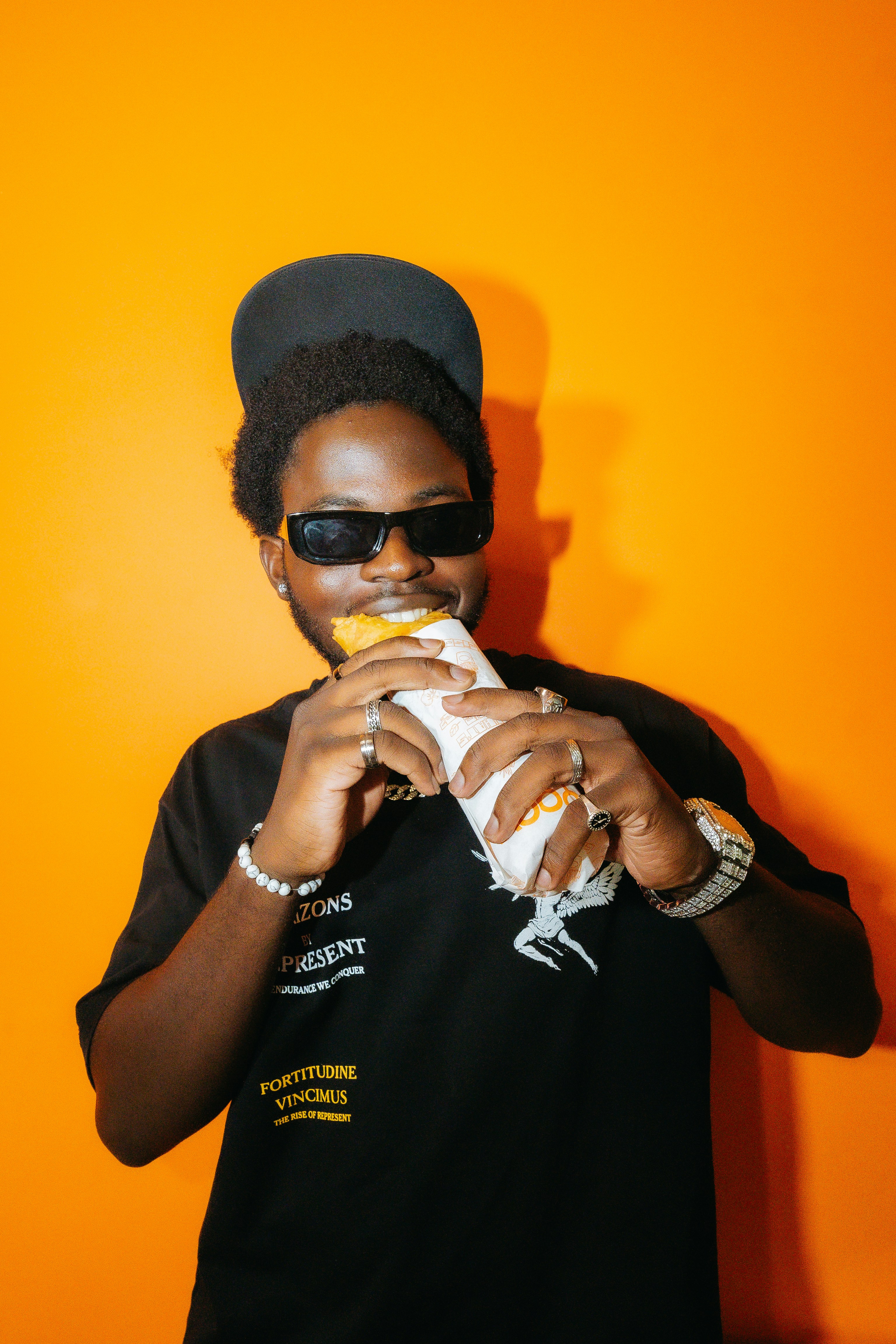 Man wearing sunglasses and cap eating a sandwich.
