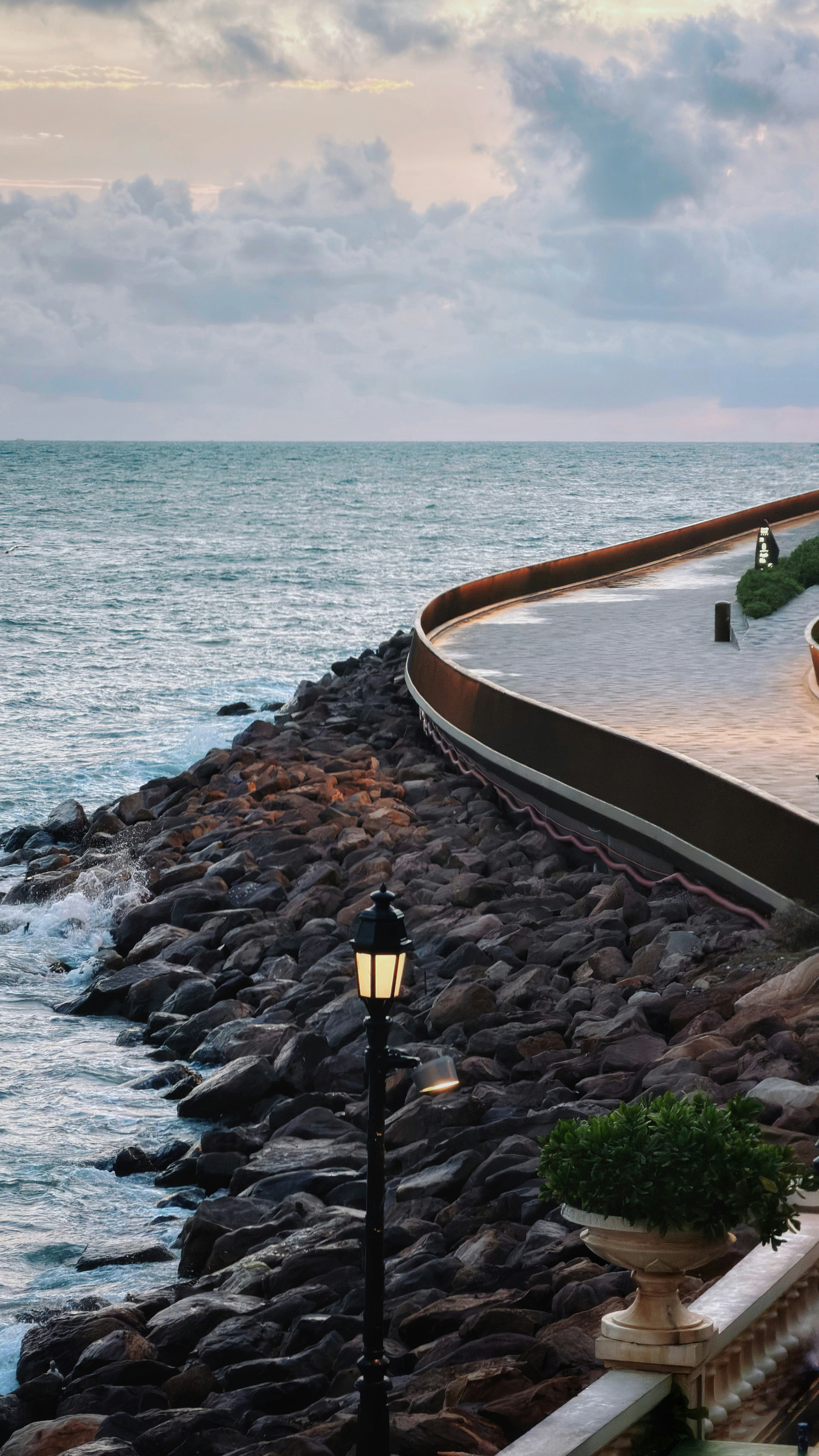 Curved ocean promenade with a lit lamppost at dusk.