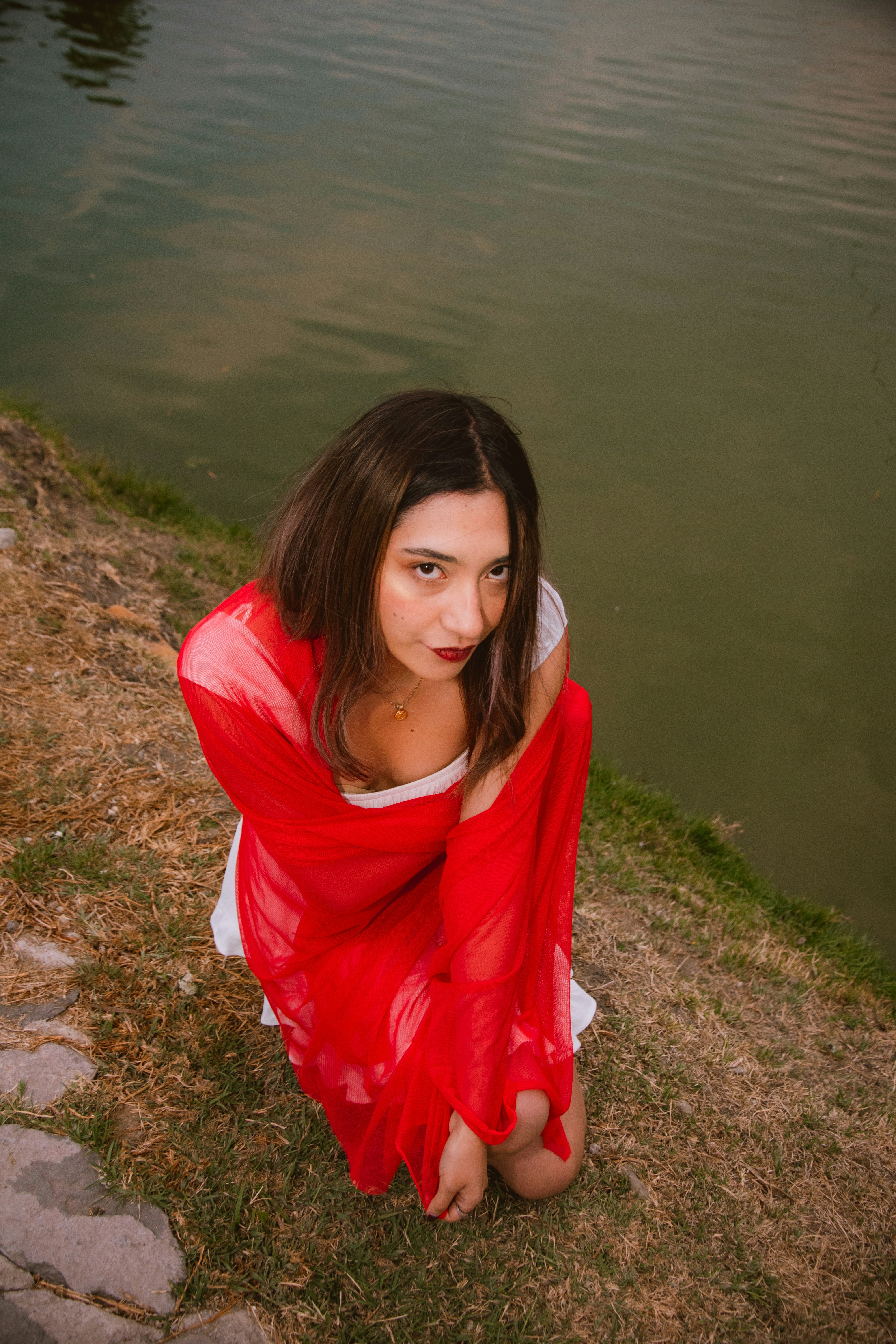 Young woman in red shawl kneeling by water, looking up