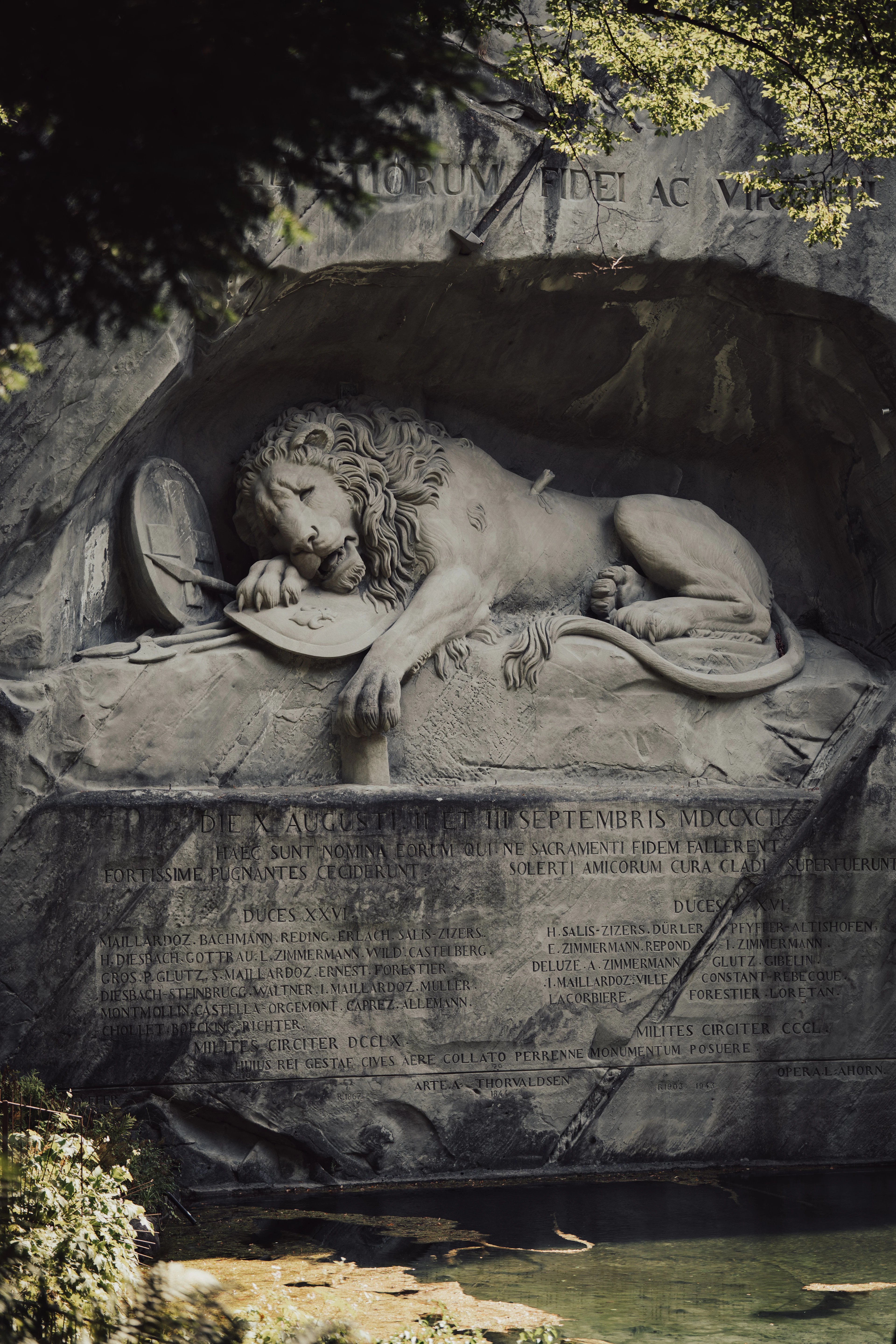 Lucerne's Lion Monument: the dying lion carved into the sandstone cliff above its pool, framed by overhanging green branches, with the Latin inscription and the roll of officers' names cut into the rock below.