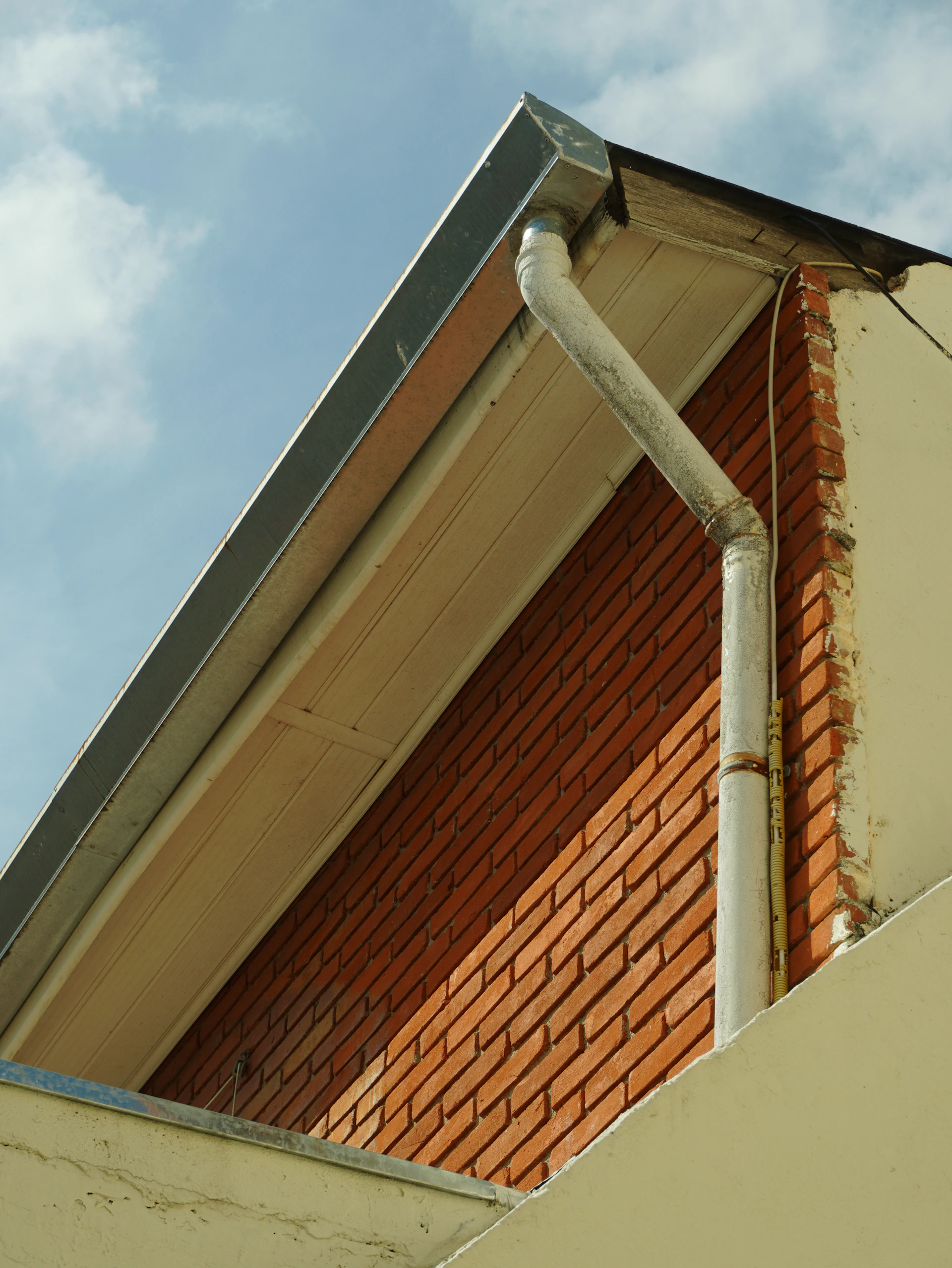A white downspout attached to a red brick wall under a roof eave
