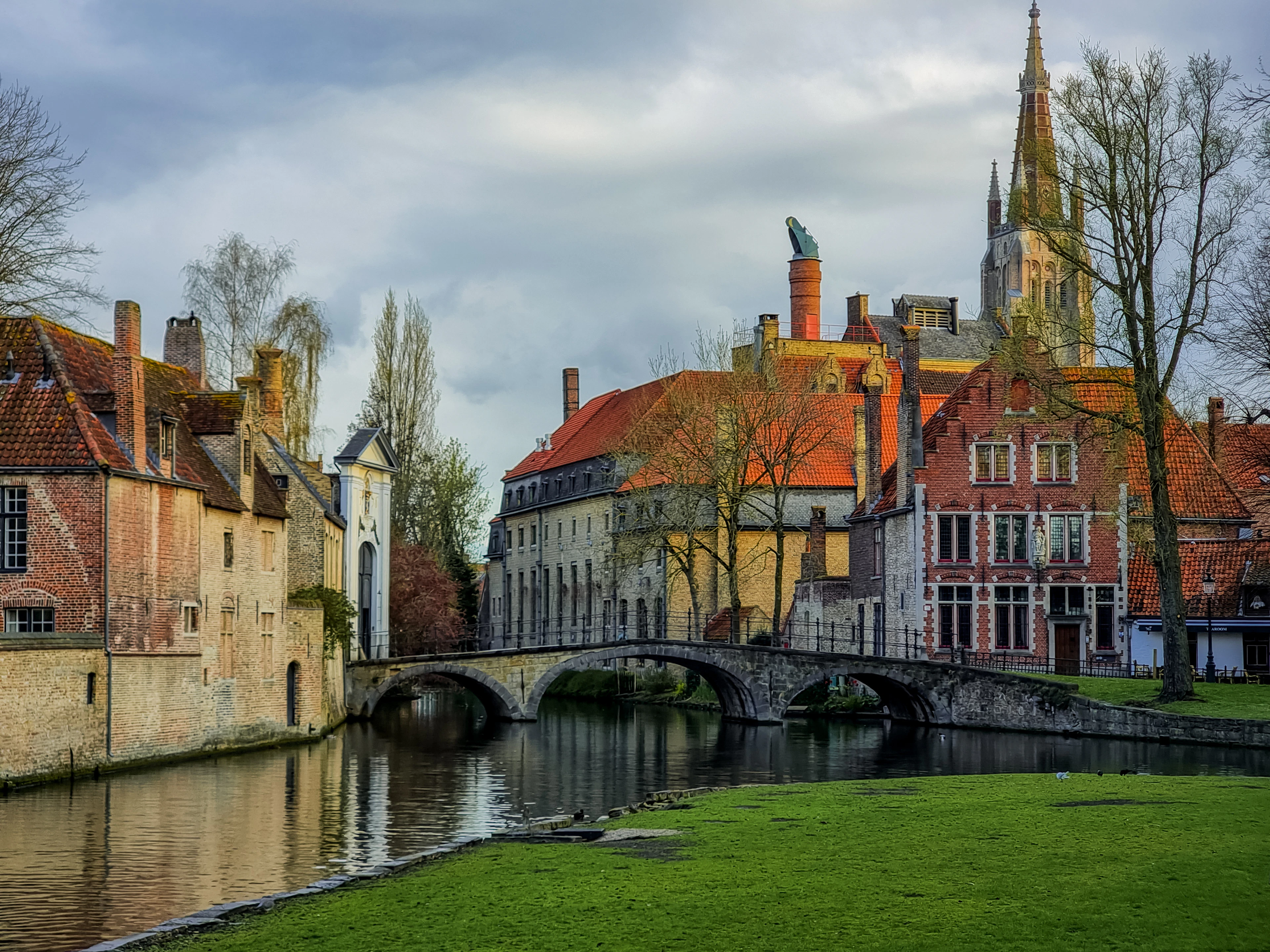 A low three-arched stone bridge over a still canal at the Begijnhof in Bruges, with lichened brick houses and orange pantile roofs on the far bank and the tall gothic spire of the Church of Our Lady rising behind under a brooding sky.
