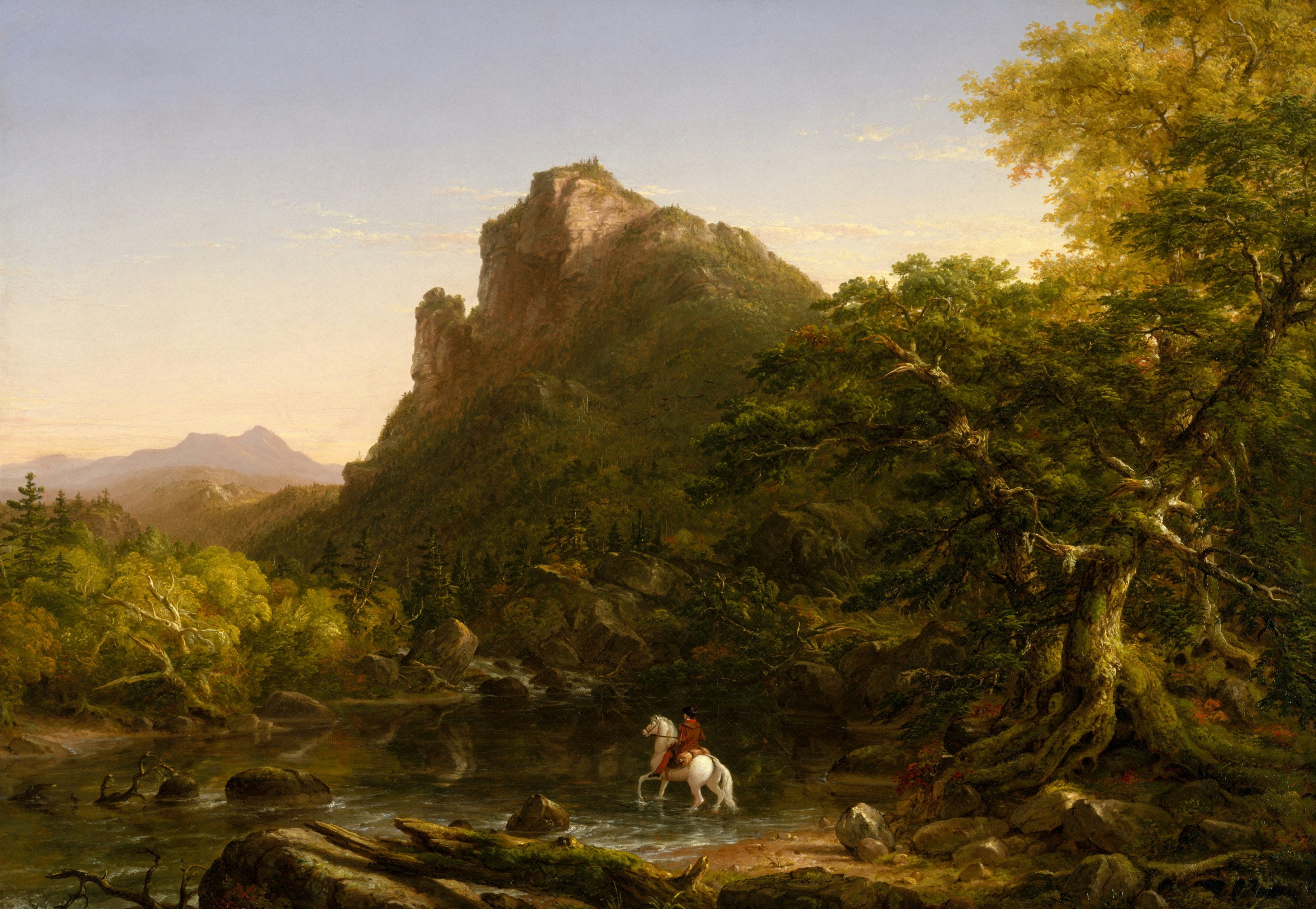 A person on a white horse crossing a river below a rocky mountain peak.