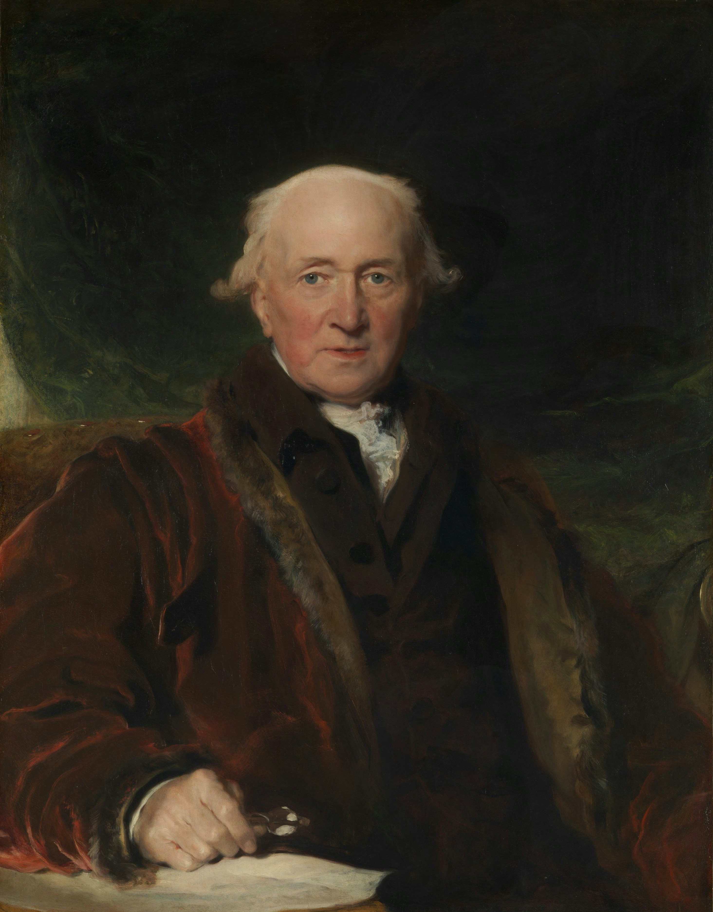 An elderly man in a dark coat with fur trim against a dark background.