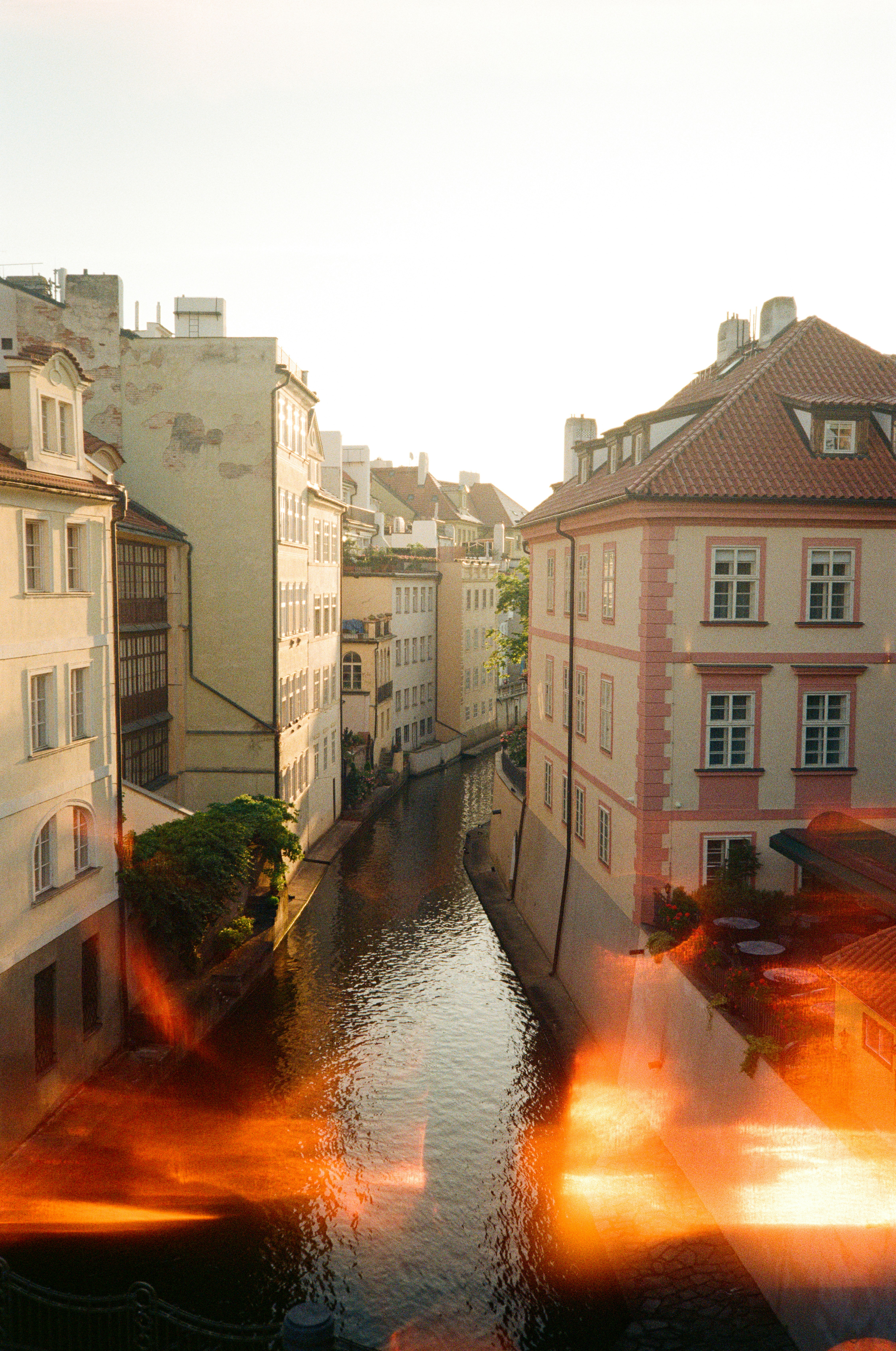 A narrow canal between historic buildings in Prague with warm light flare