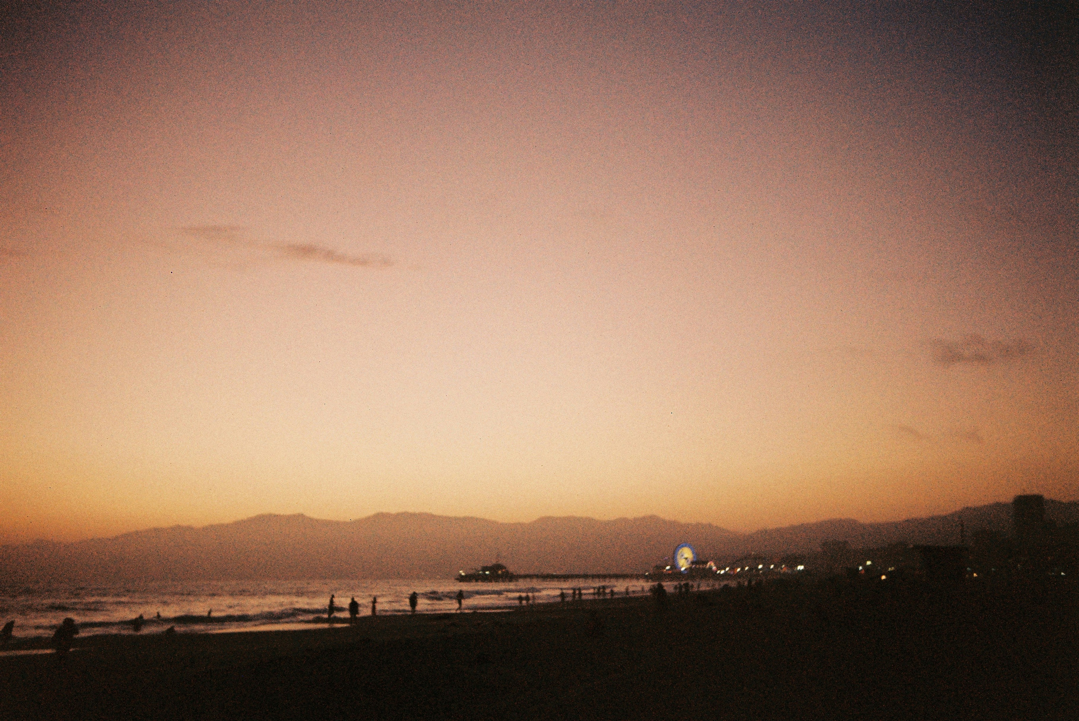 Silhouettes of people on a beach with a pier and ferris wheel at low sun.