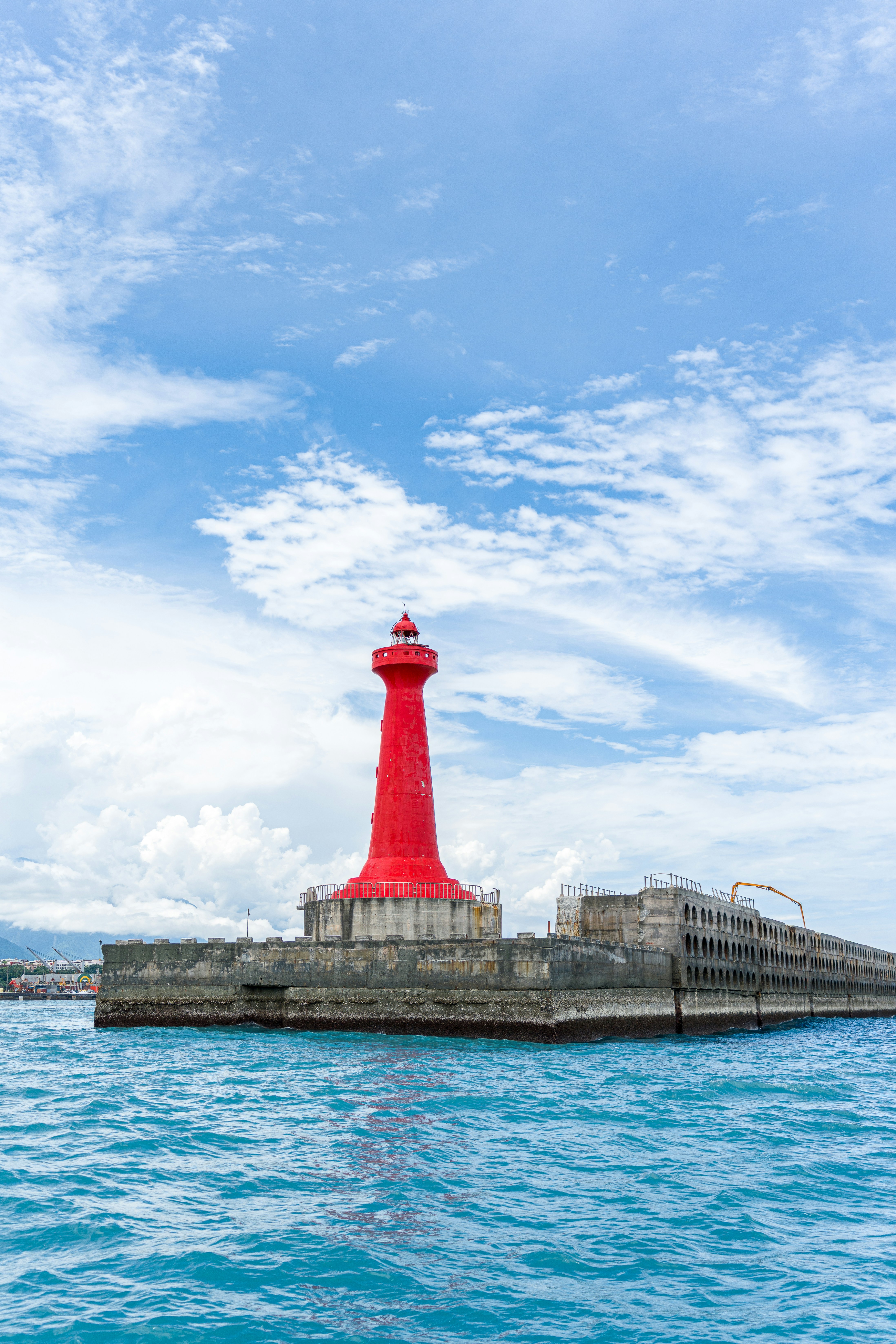 A bright red lighthouse on a concrete pier surrounded by blue ocean water.