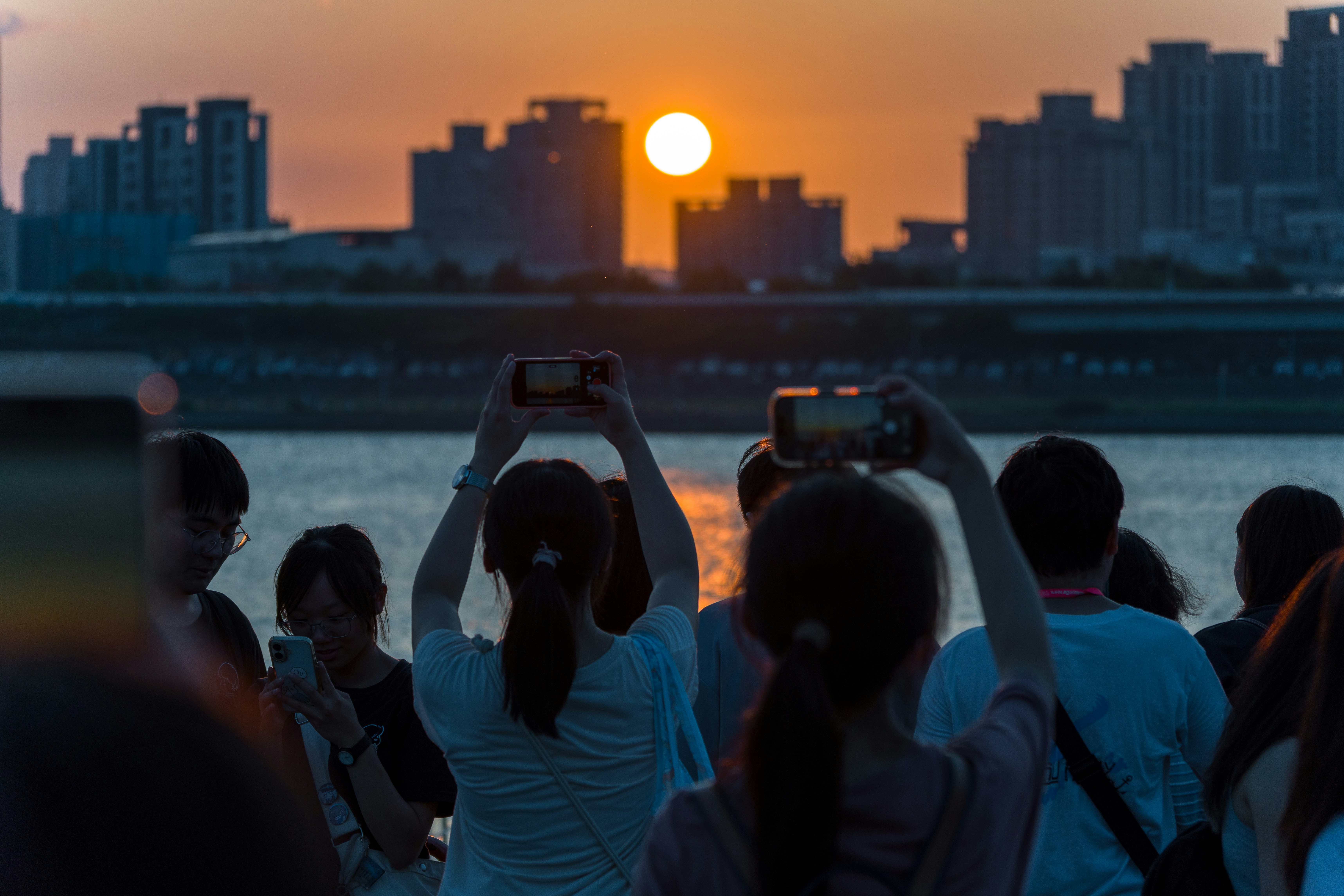 A group of people holding up smartphones to photograph a low sun over city buildings.