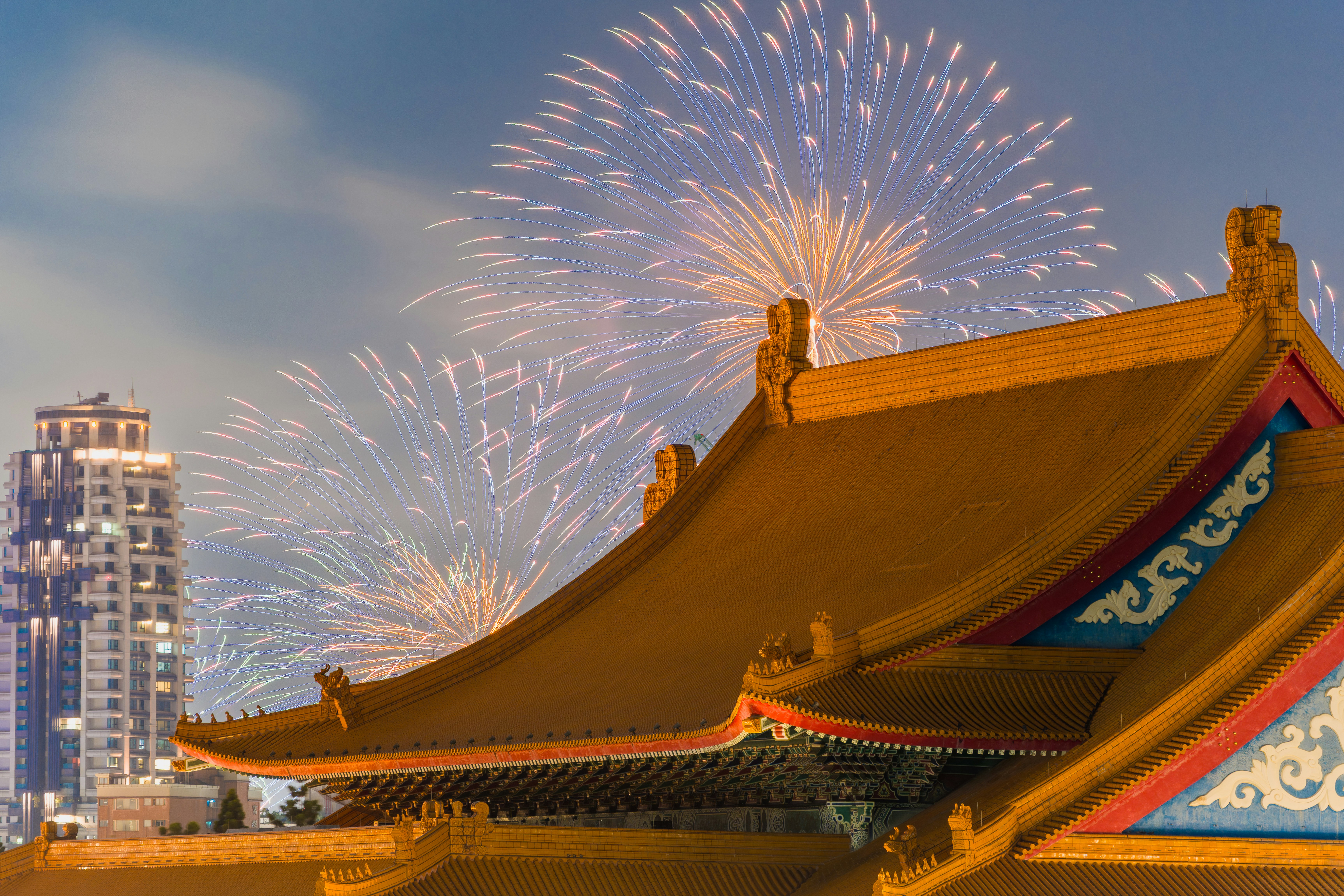 Fireworks exploding above a traditional Chinese temple roof with a modern skyscraper nearby.