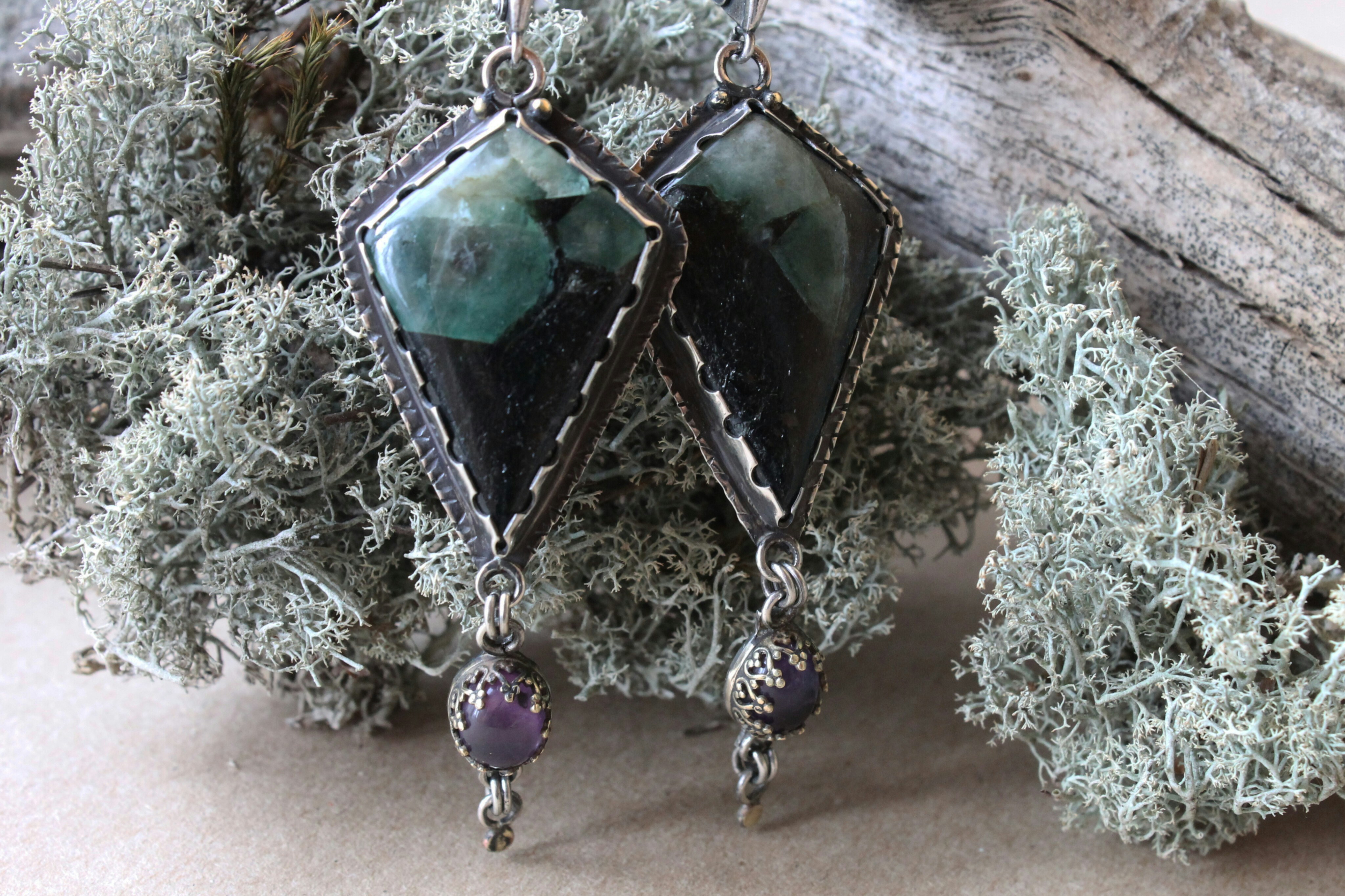 Deux boucles d’oreilles en forme de cerf-volant avec des pierres vertes et noires suspendues au-dessus d’un lichen gris