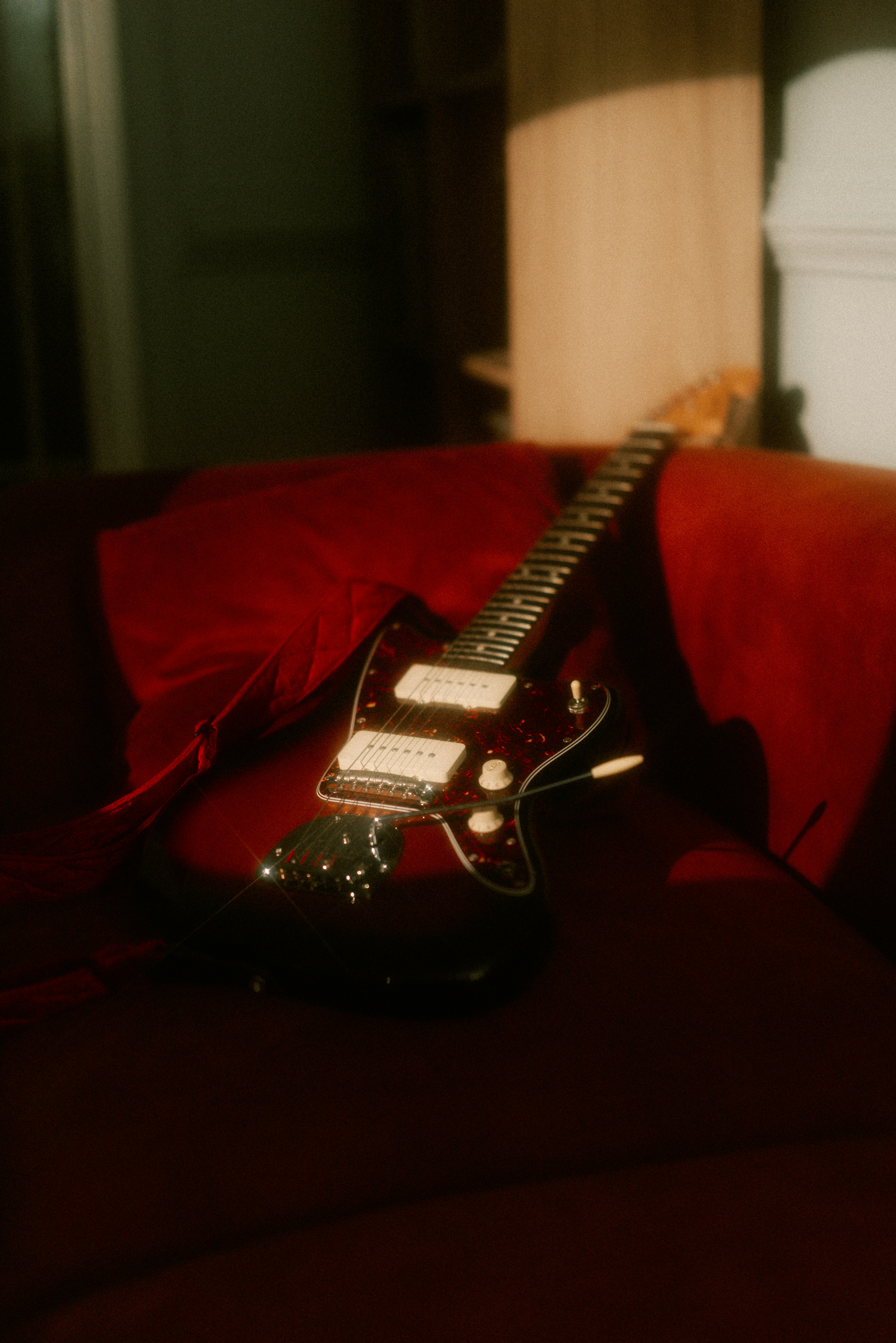 An electric guitar with a tortoiseshell pickguard resting on a red velvet sofa.