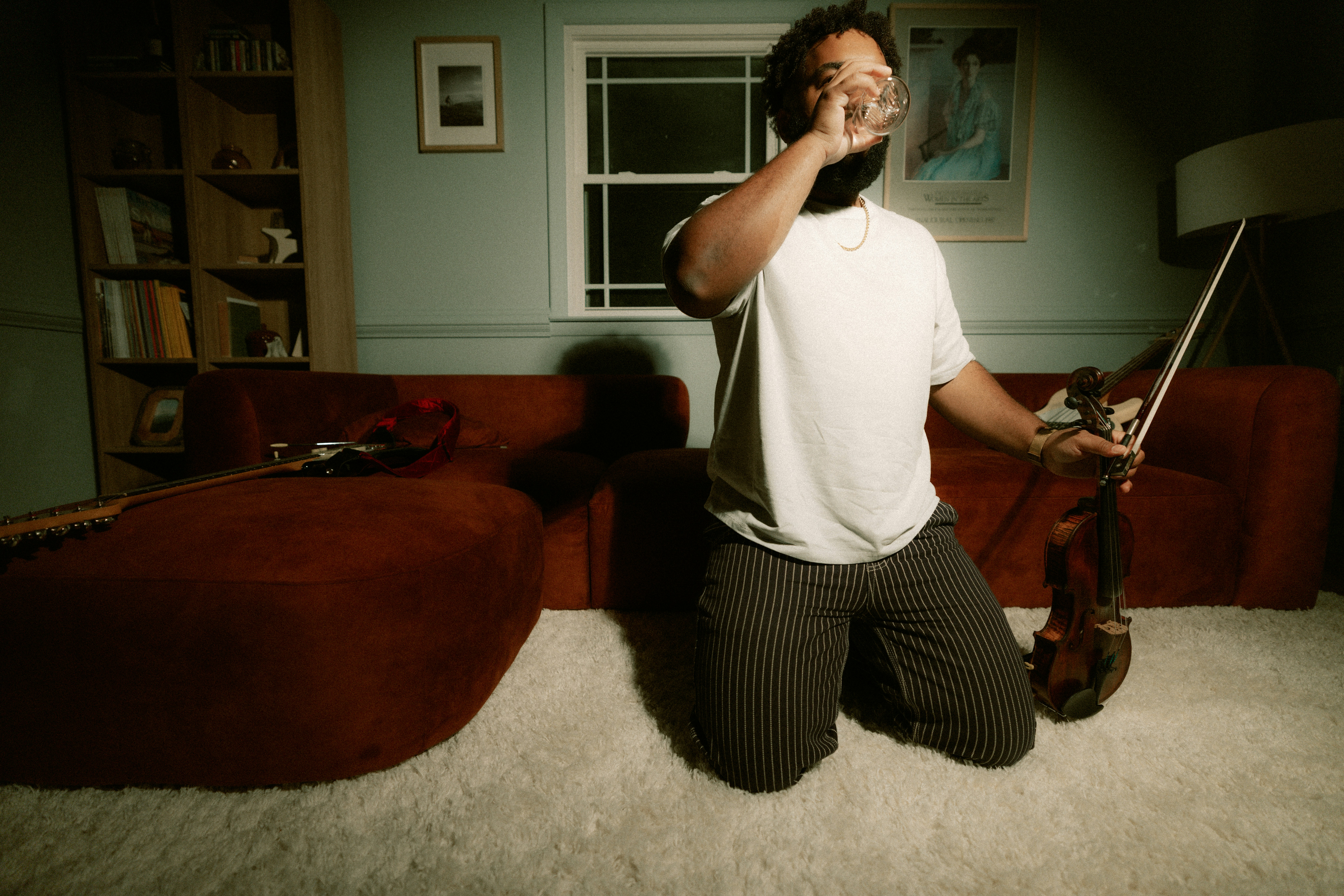 A person kneeling on a rug, drinking from a glass while holding a violin.