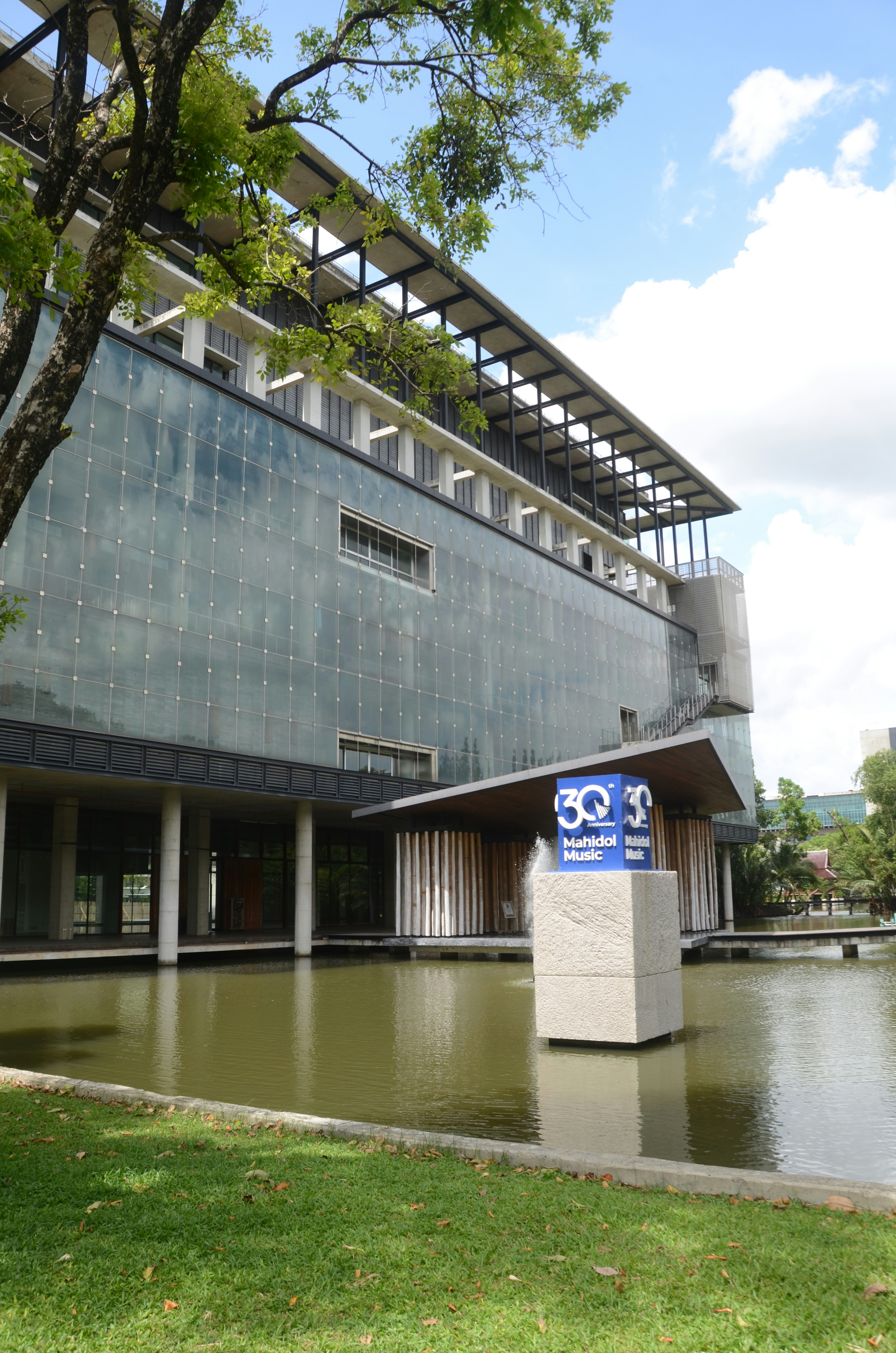 Un moderno edificio di vetro con una fontana e un'insegna Mahidol Music accanto a uno stagno.