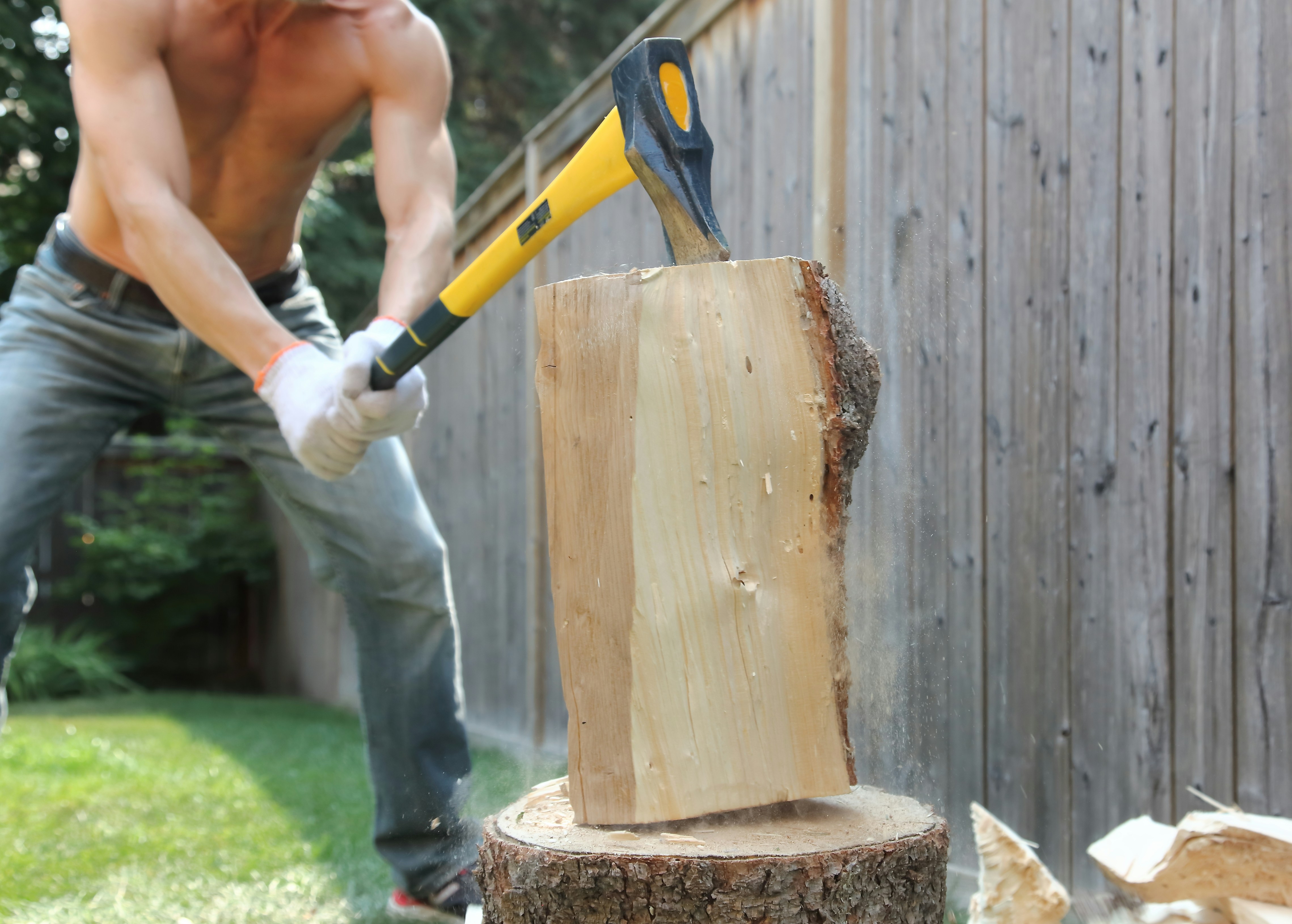 A shirtless person splitting a log with a yellow axe in a backyard.
