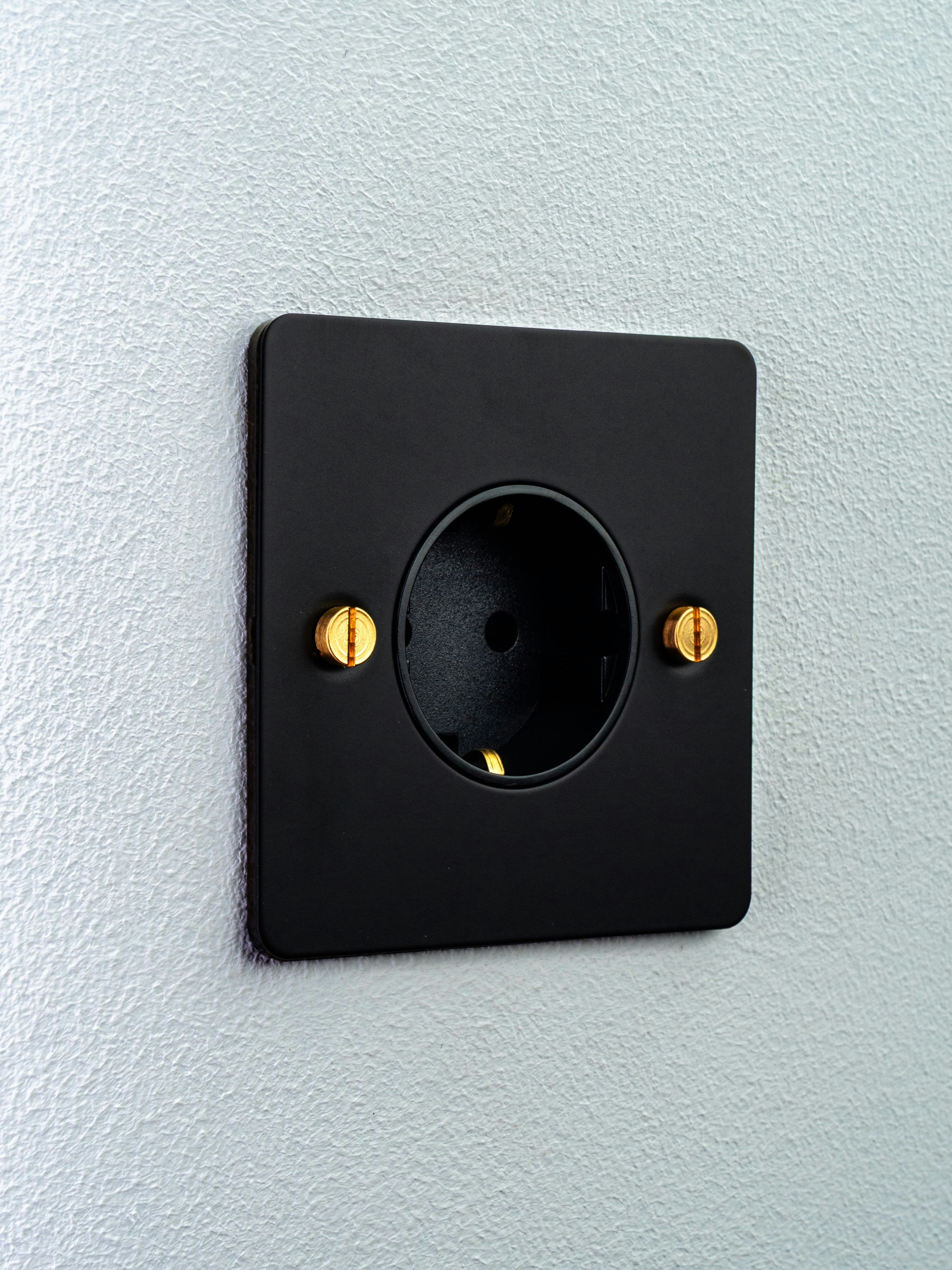 A matte black electrical outlet with brass screws on a textured white wall
