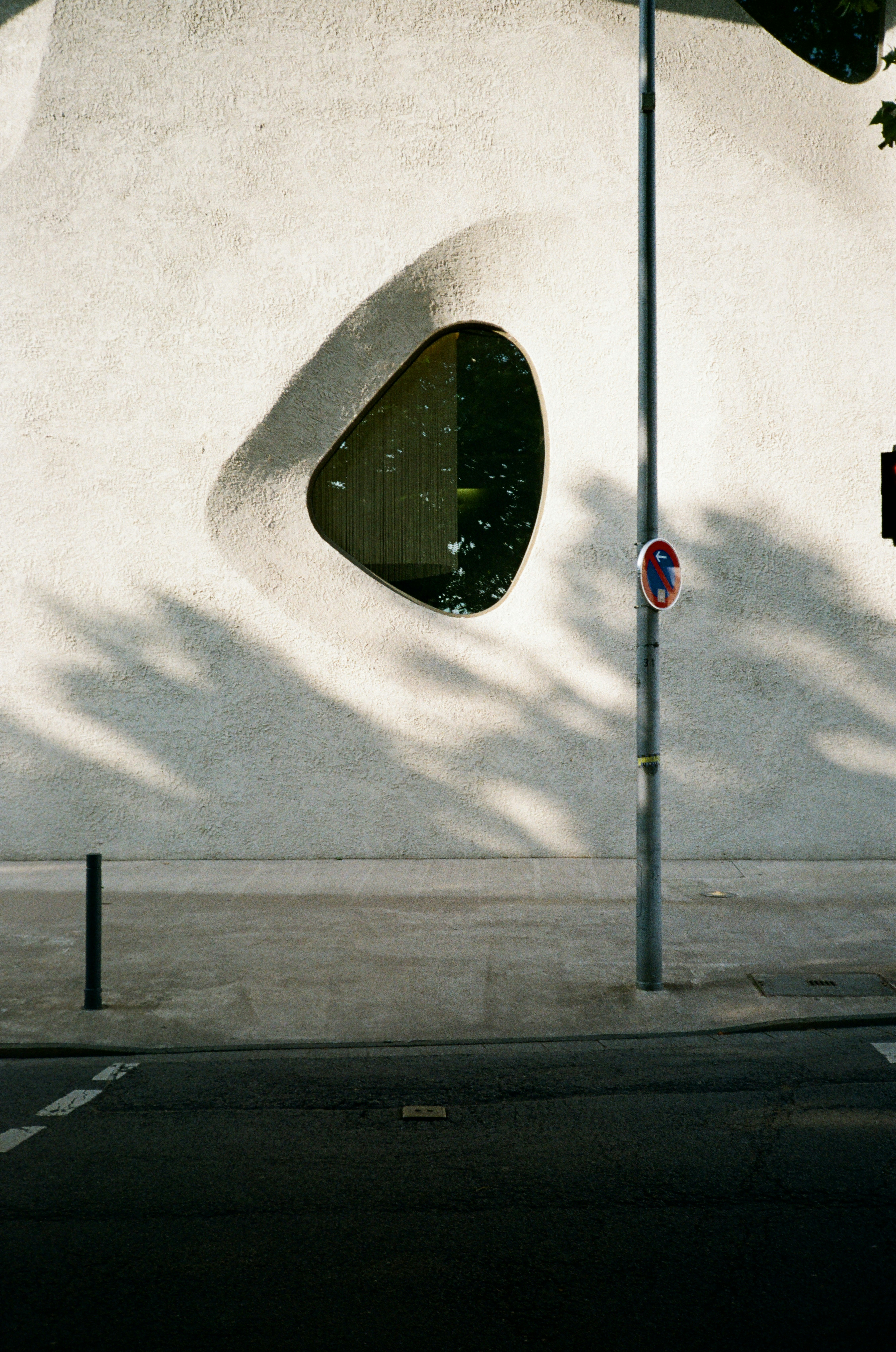 A textured white wall with an organic shaped window above a city sidewalk.