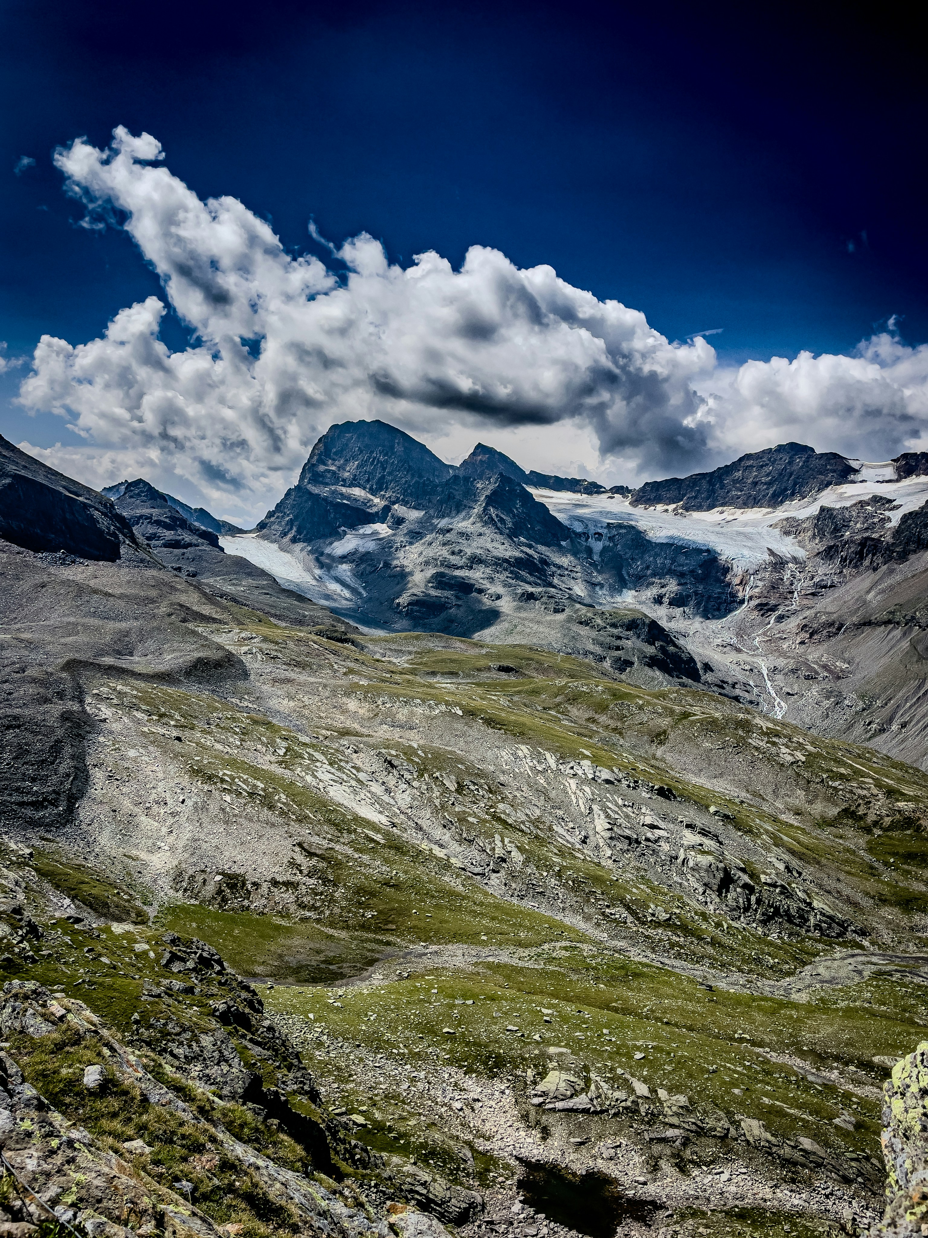 Rocky mountain peaks with glaciers above a green alpine valley under a cloudy sky.
