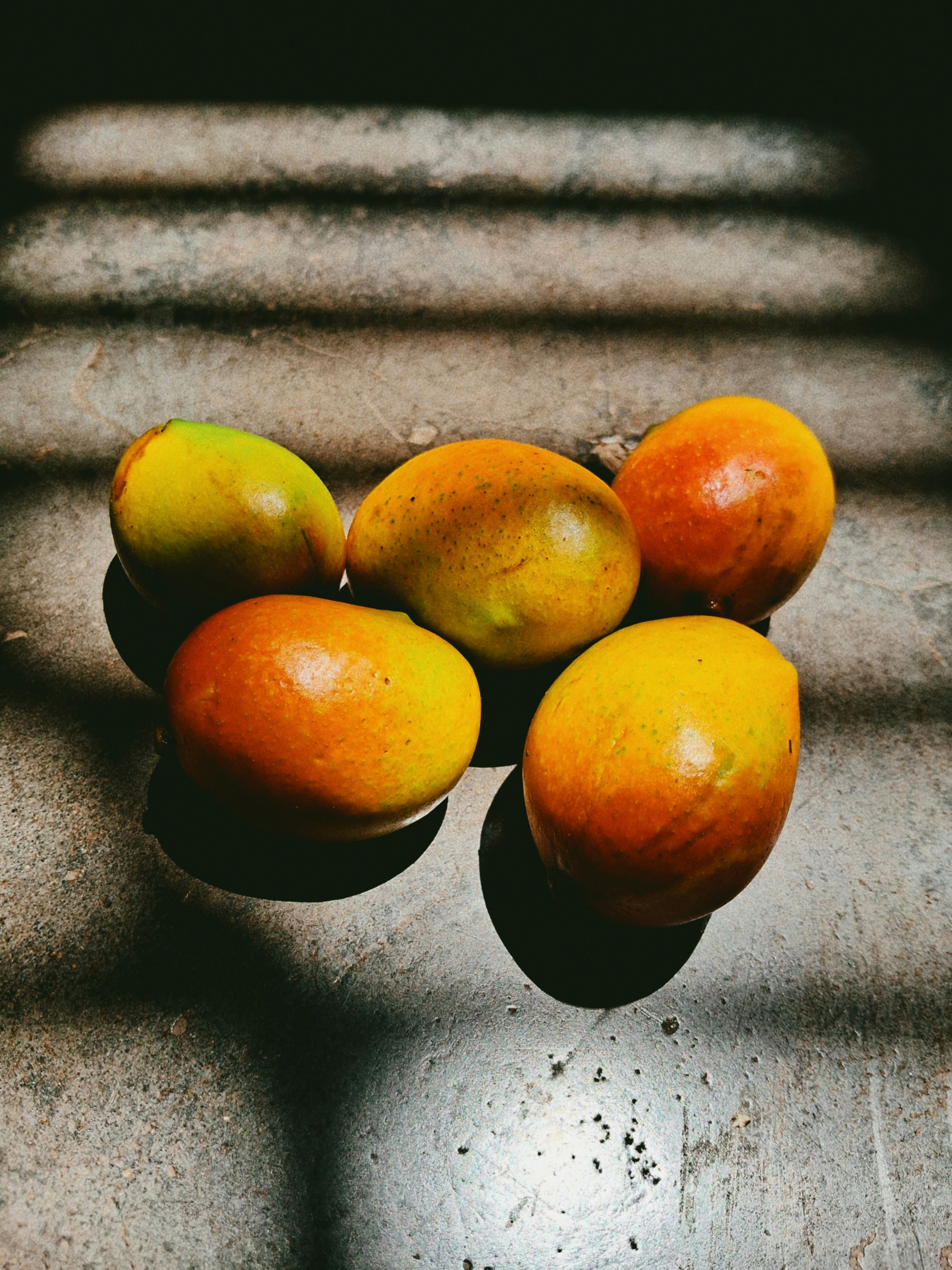 Five mangoes with yellow and orange skin on a textured grey surface