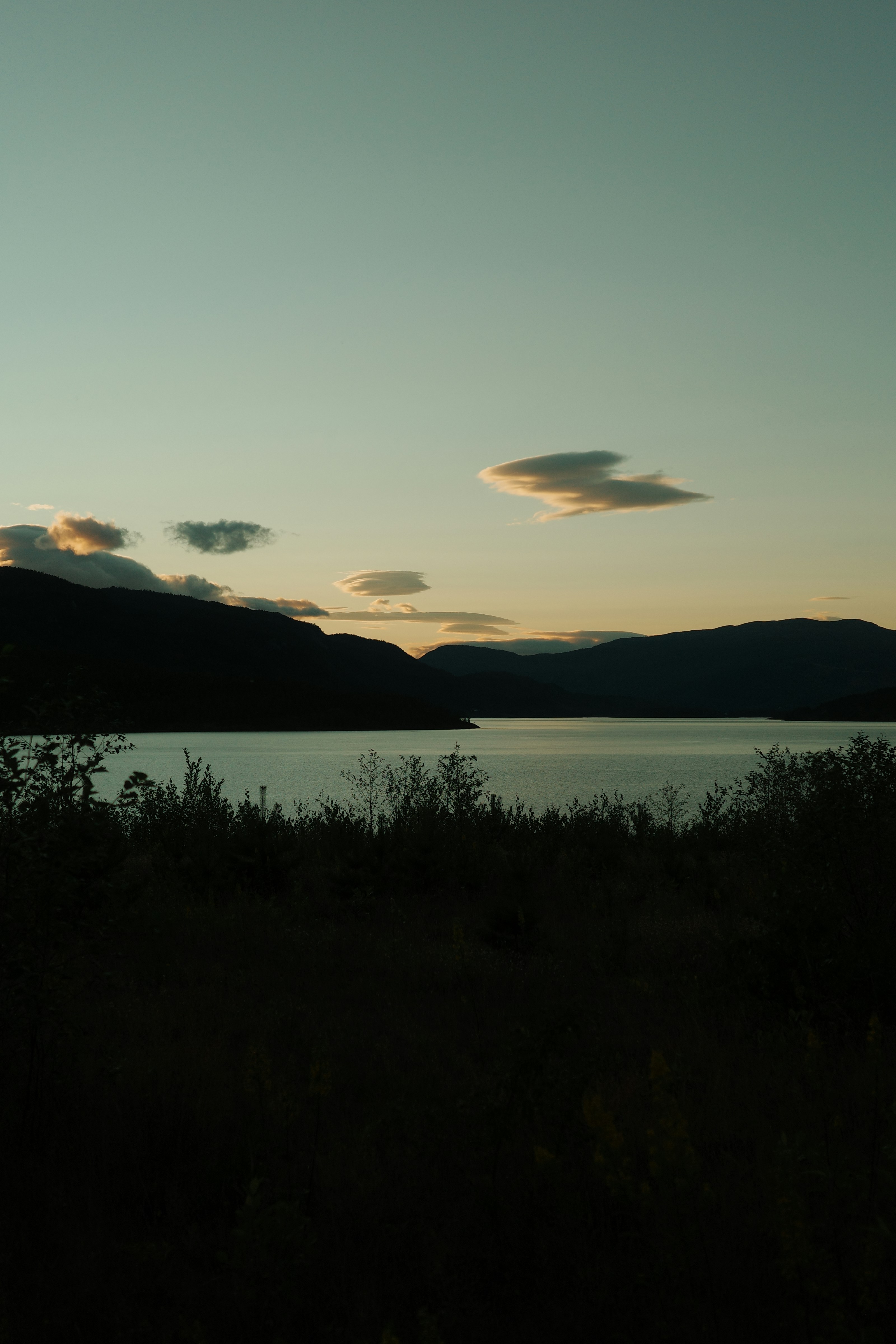 Linsenartige Wolken über einem dunklen See und silhouettierten Berge unter einem blassen Himmel