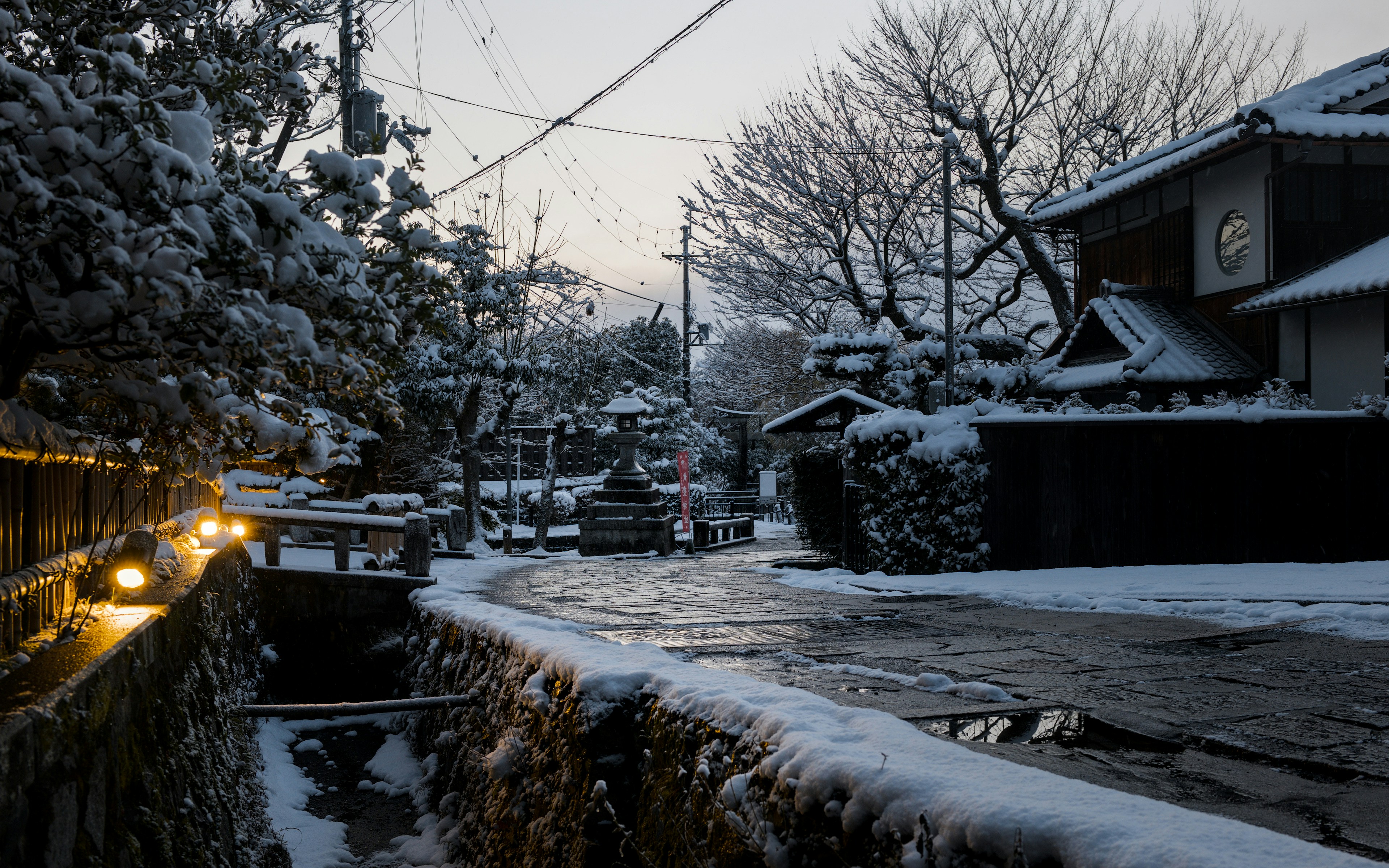 Un chemin de pierre enneigé au bord d’un canal avec des lumières brillantes dans un village japonais.