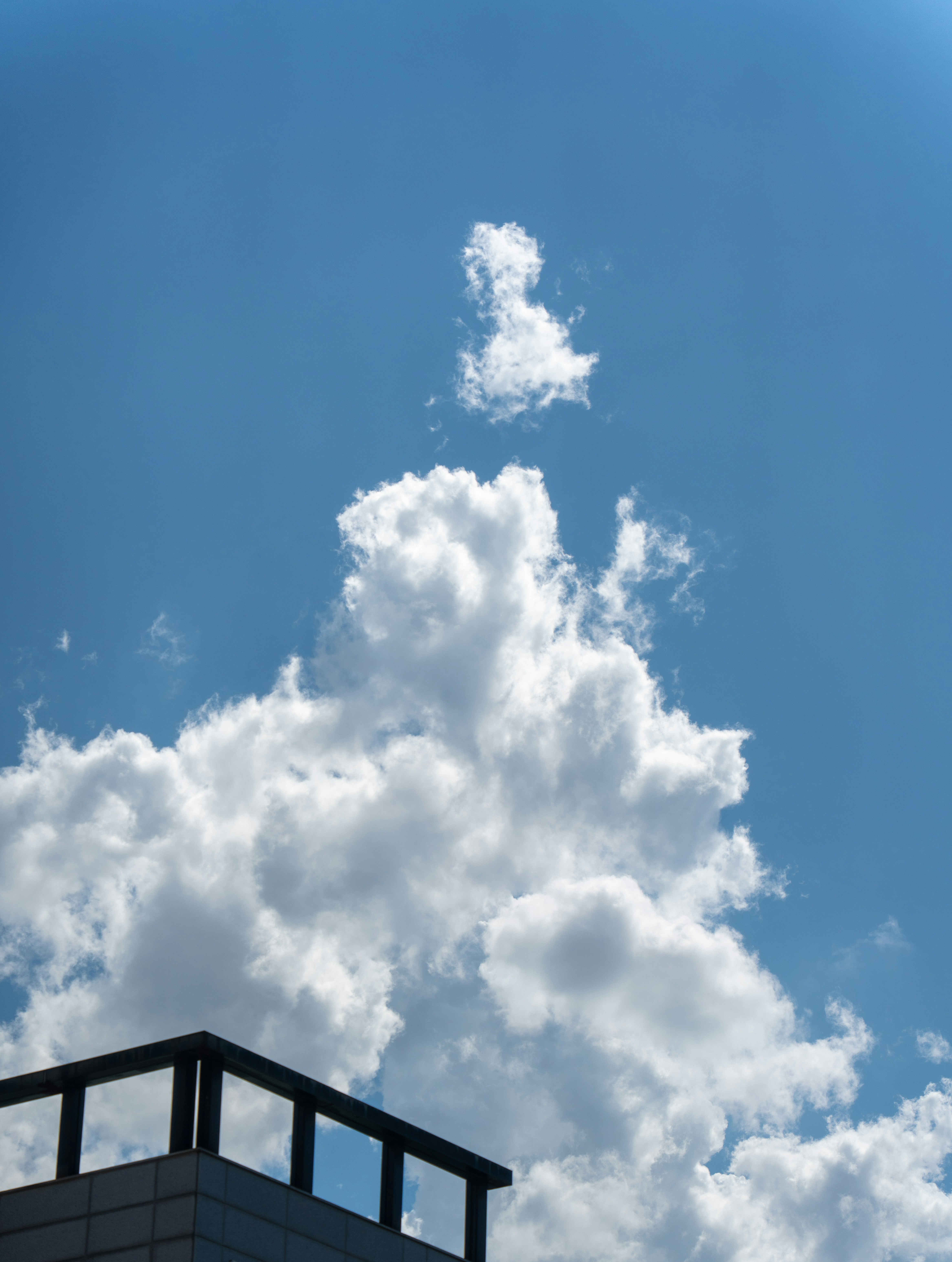 White cumulus clouds in a bright blue sky above a building rooftop.