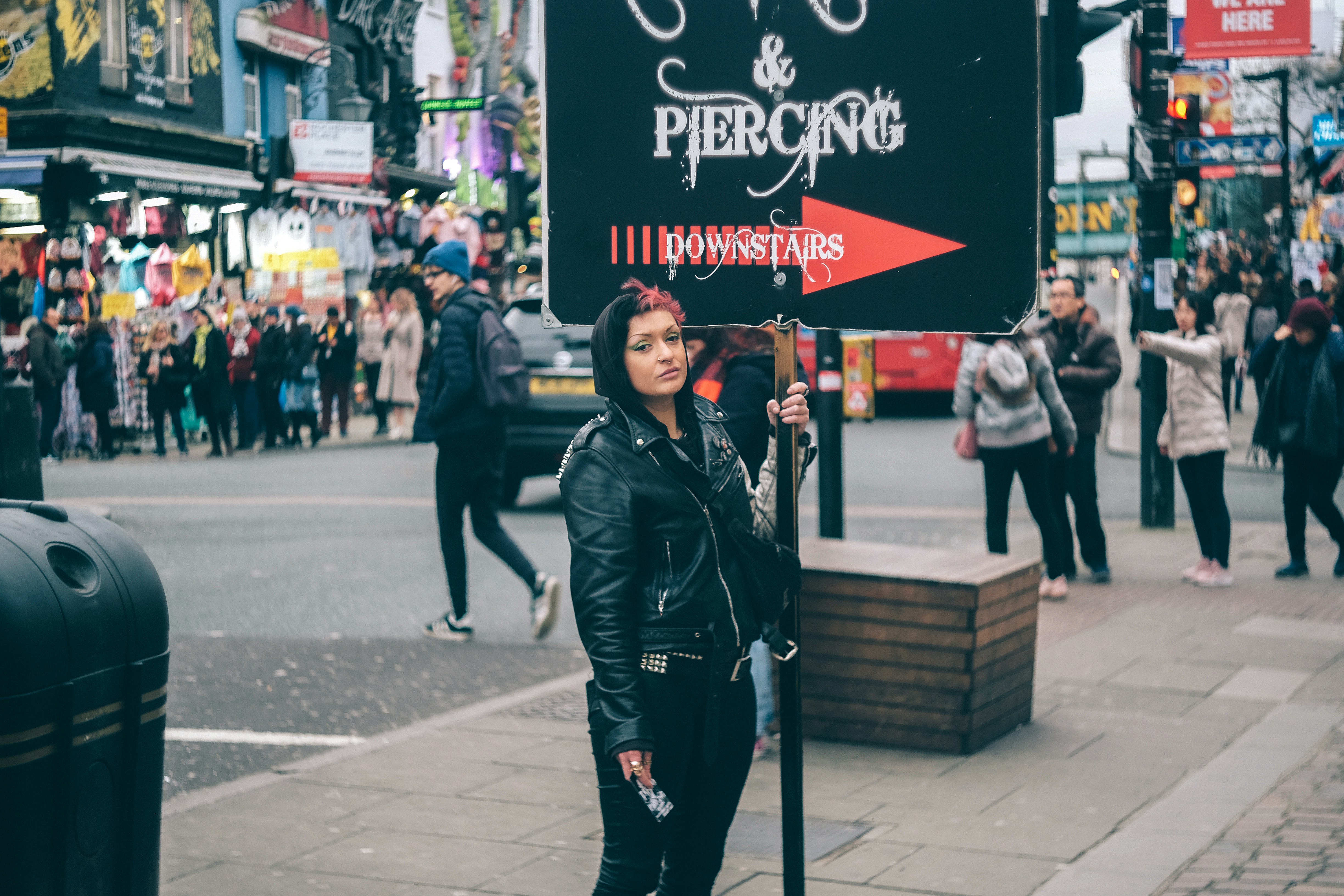 A person in a leather jacket holding a sign reading "PIERCING" on a city street.