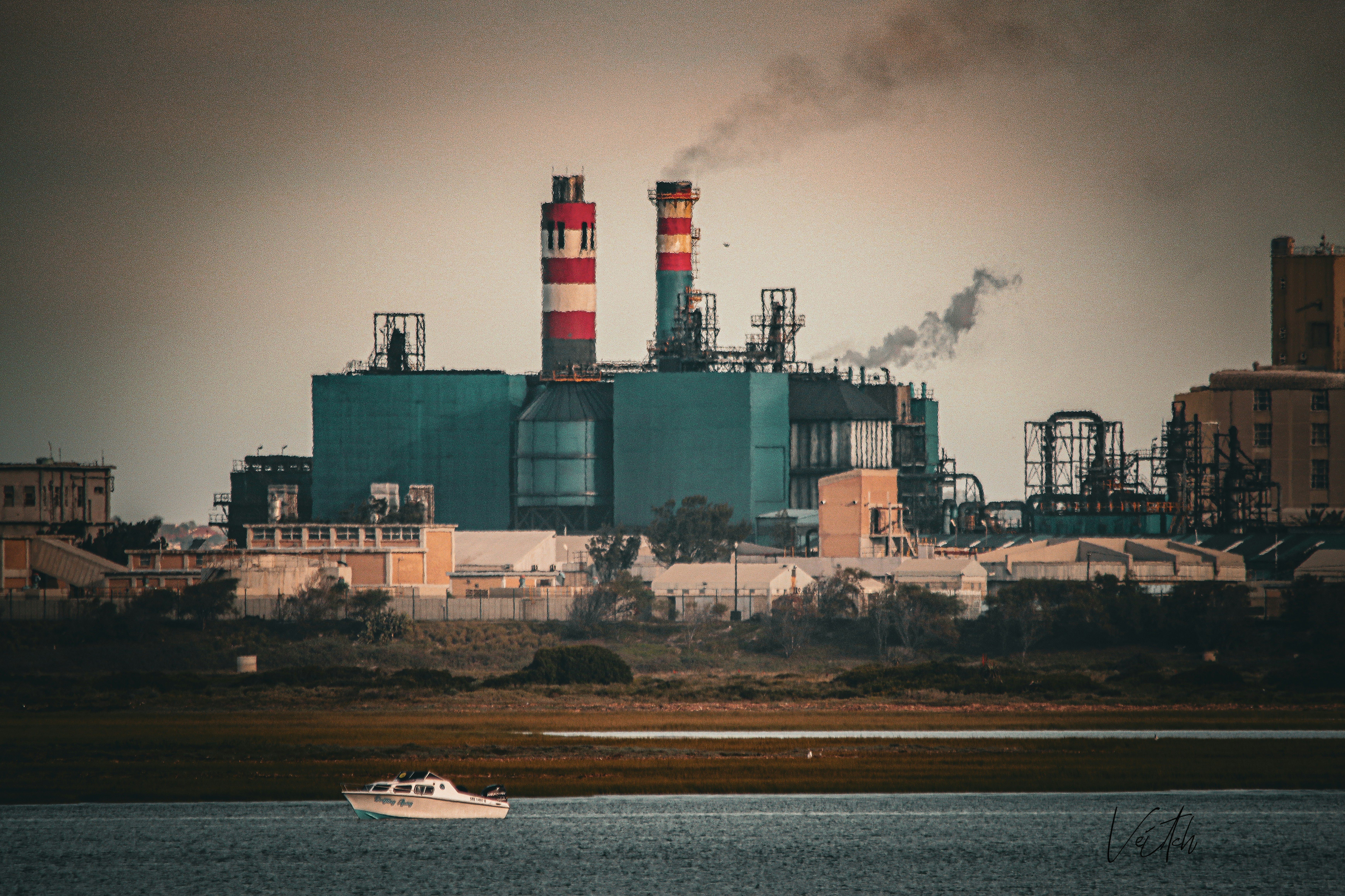A small boat on water before a large industrial factory with smoking chimneys