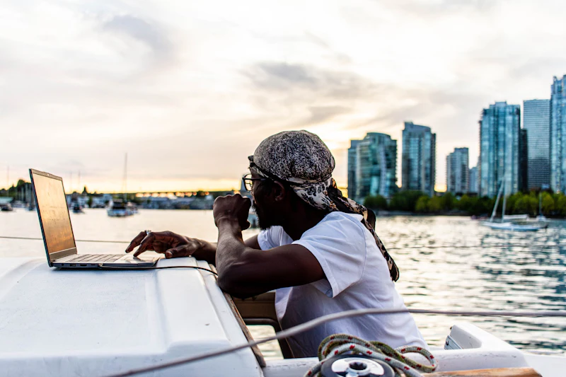 Traveller working on a laptop aboard a boat in a city harbour at sunset, connected through a phone hotspot