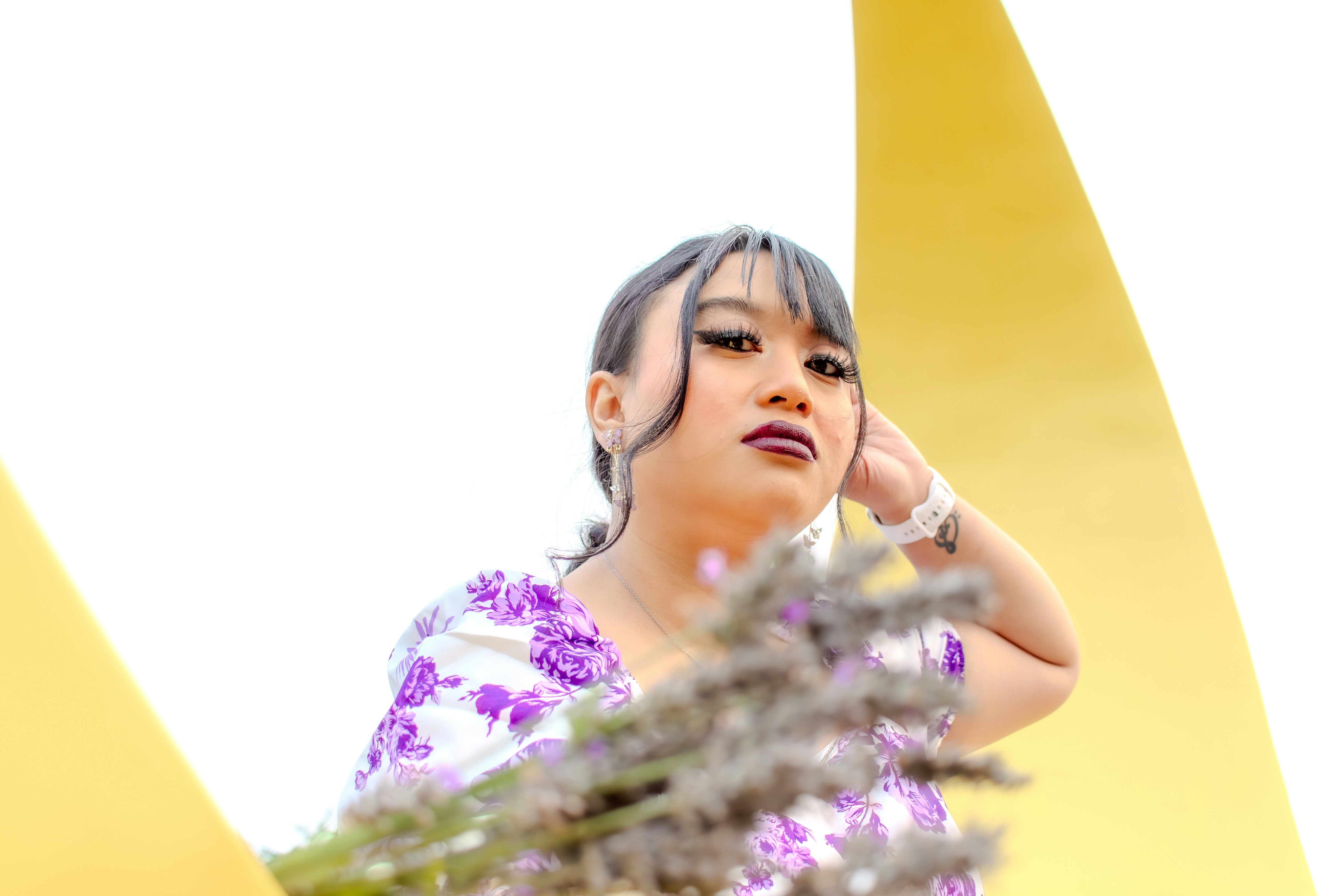 紫色の花柄トップスを着て、明るい黄色の背景にラベンダーを手に持つ女性。