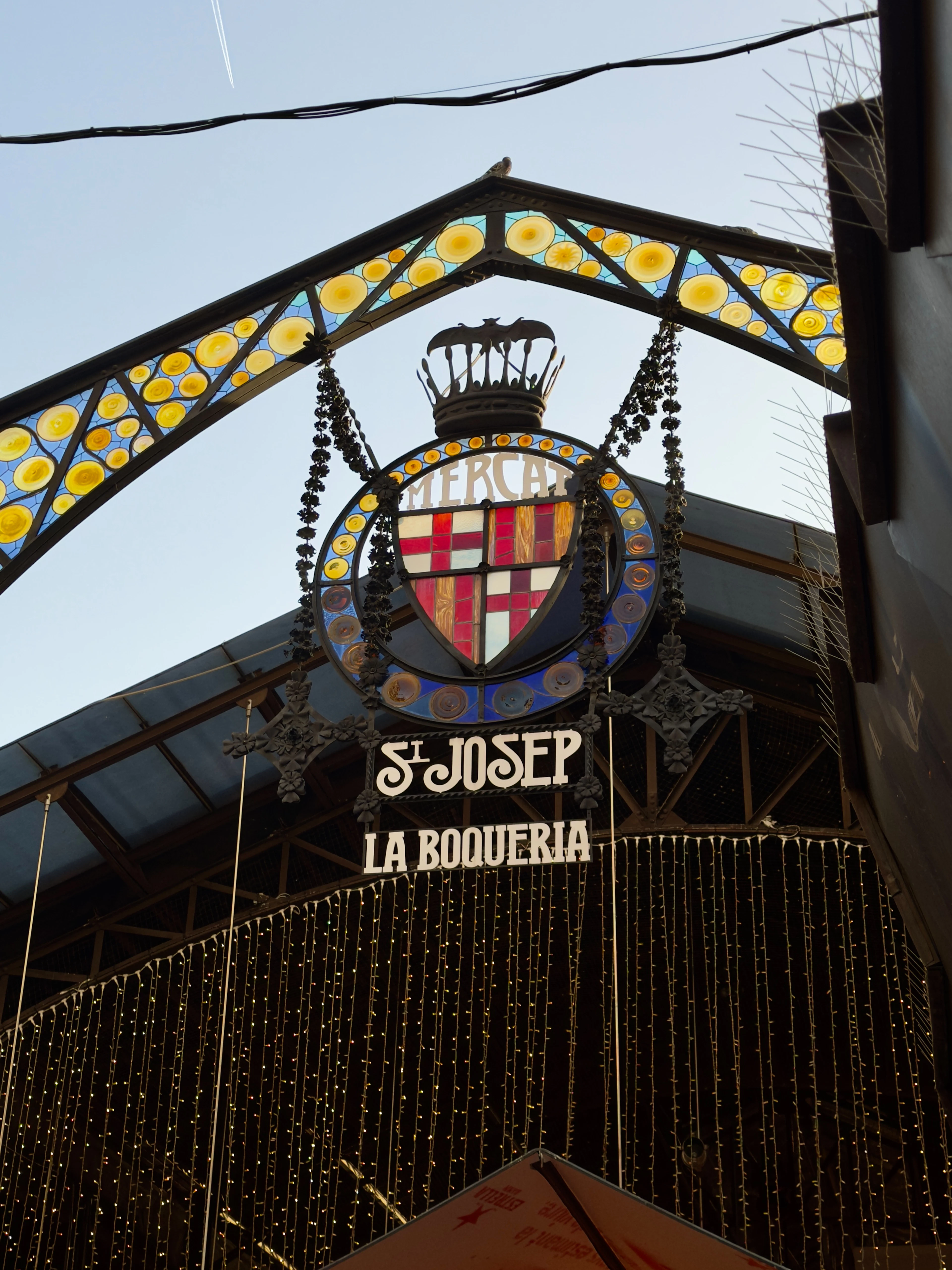 La Boqueria's ornate market archway lit by strings of bulbs at dusk.