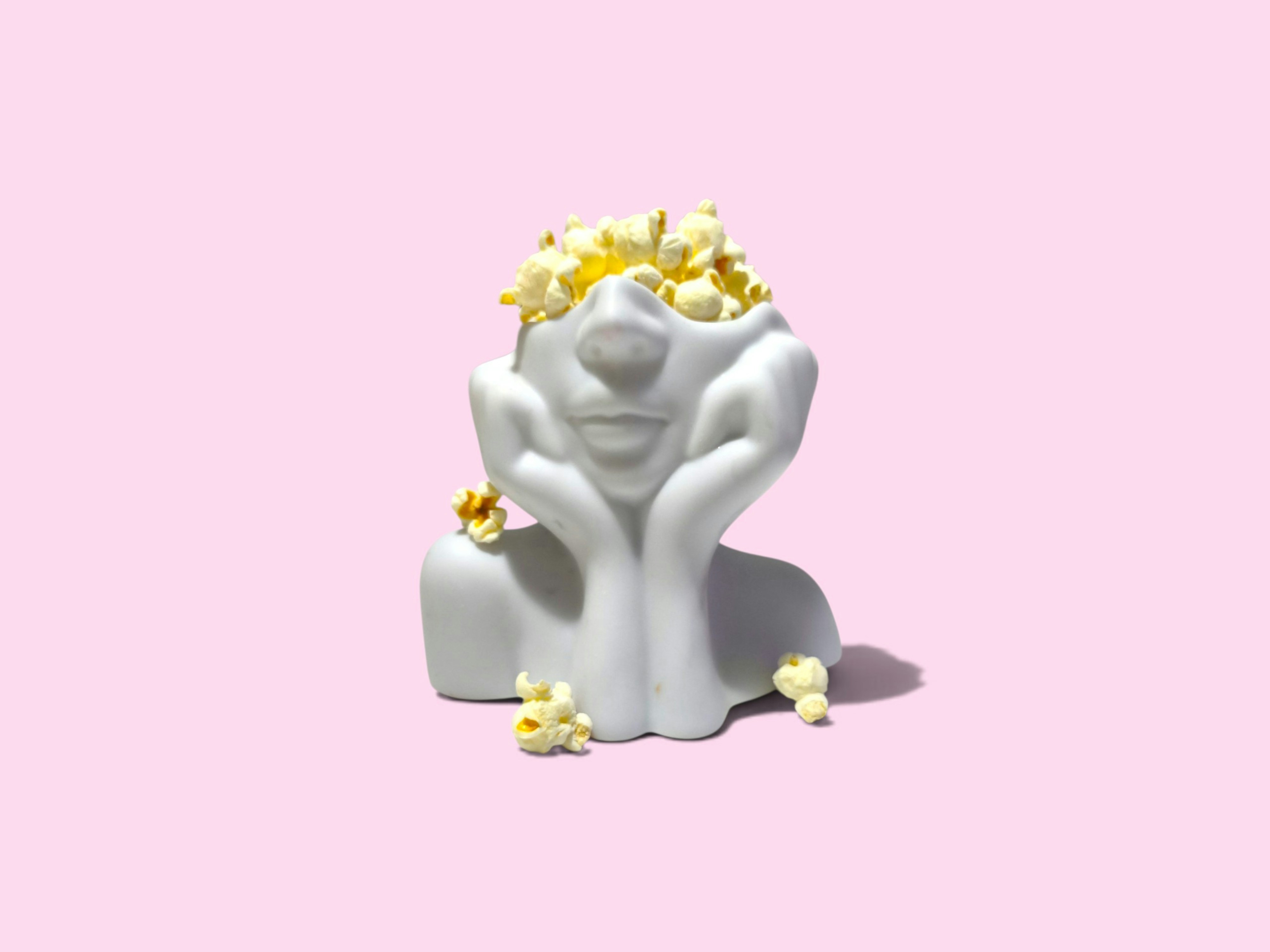 Creative surreal concept featuring a white ceramic face sculpture filled with popcorn on a pastel pink background.