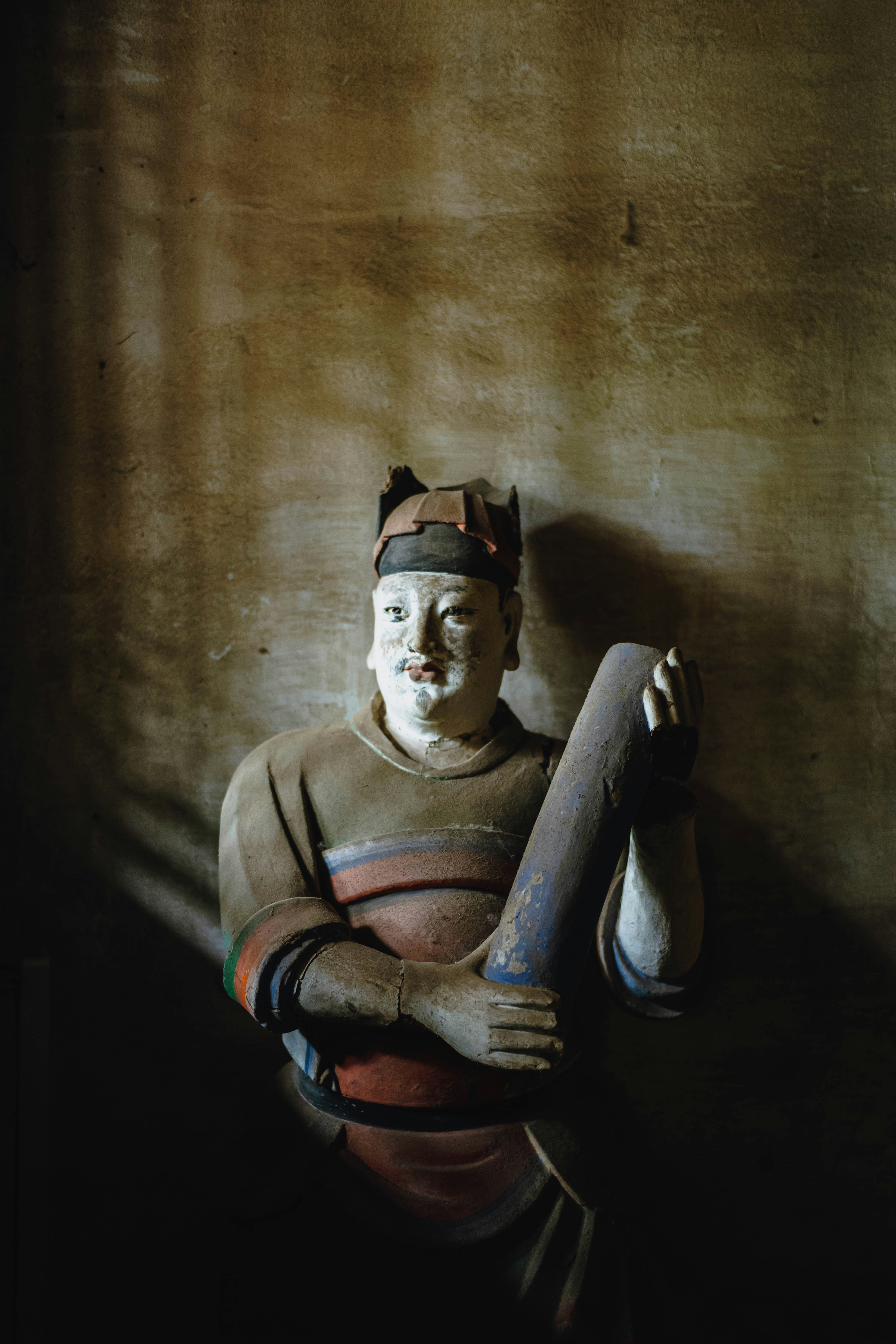 A weathered wooden statue of a person holding a blue cylinder against a wall