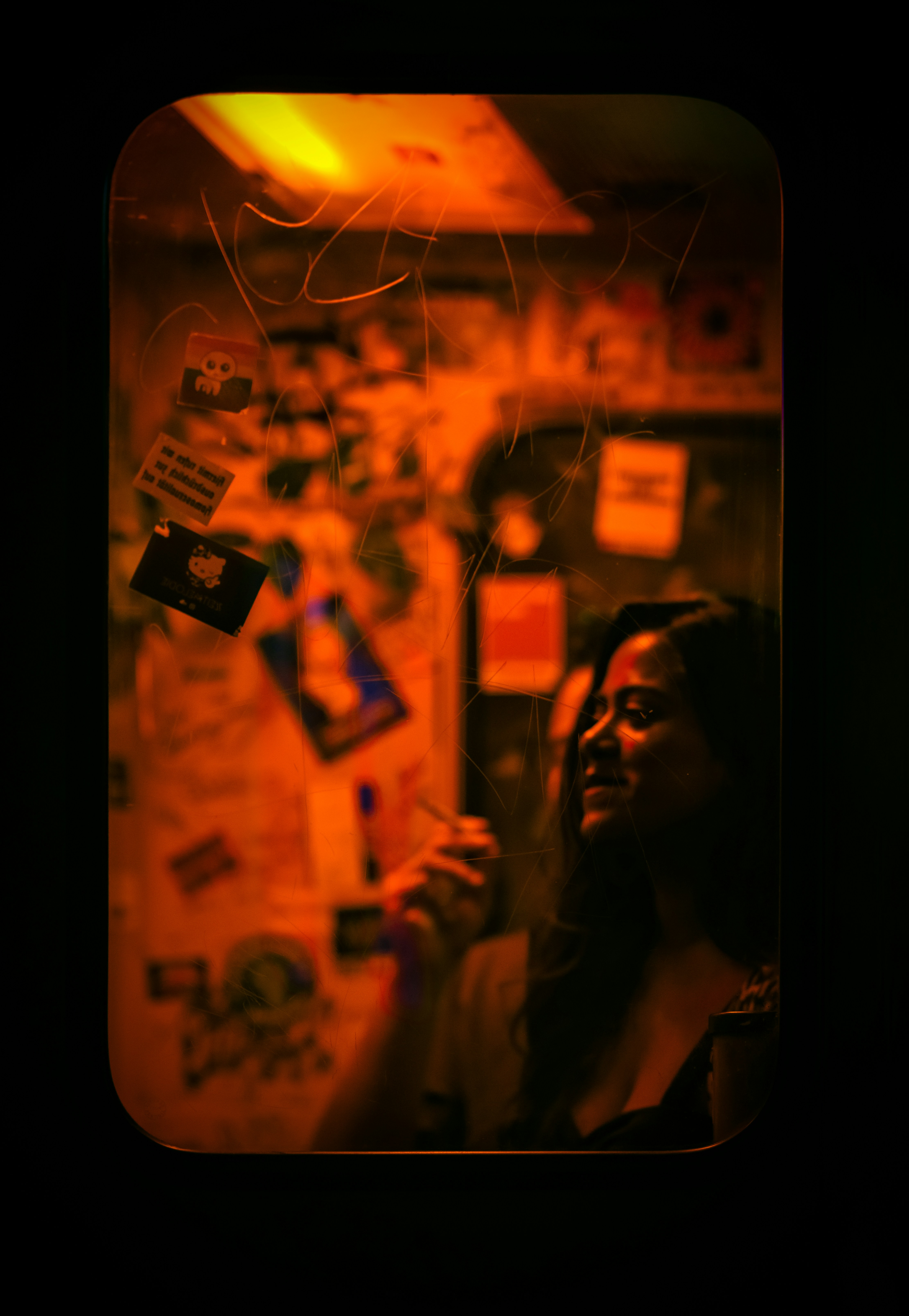 A woman reflected in a scratched mirror covered in stickers under warm orange light