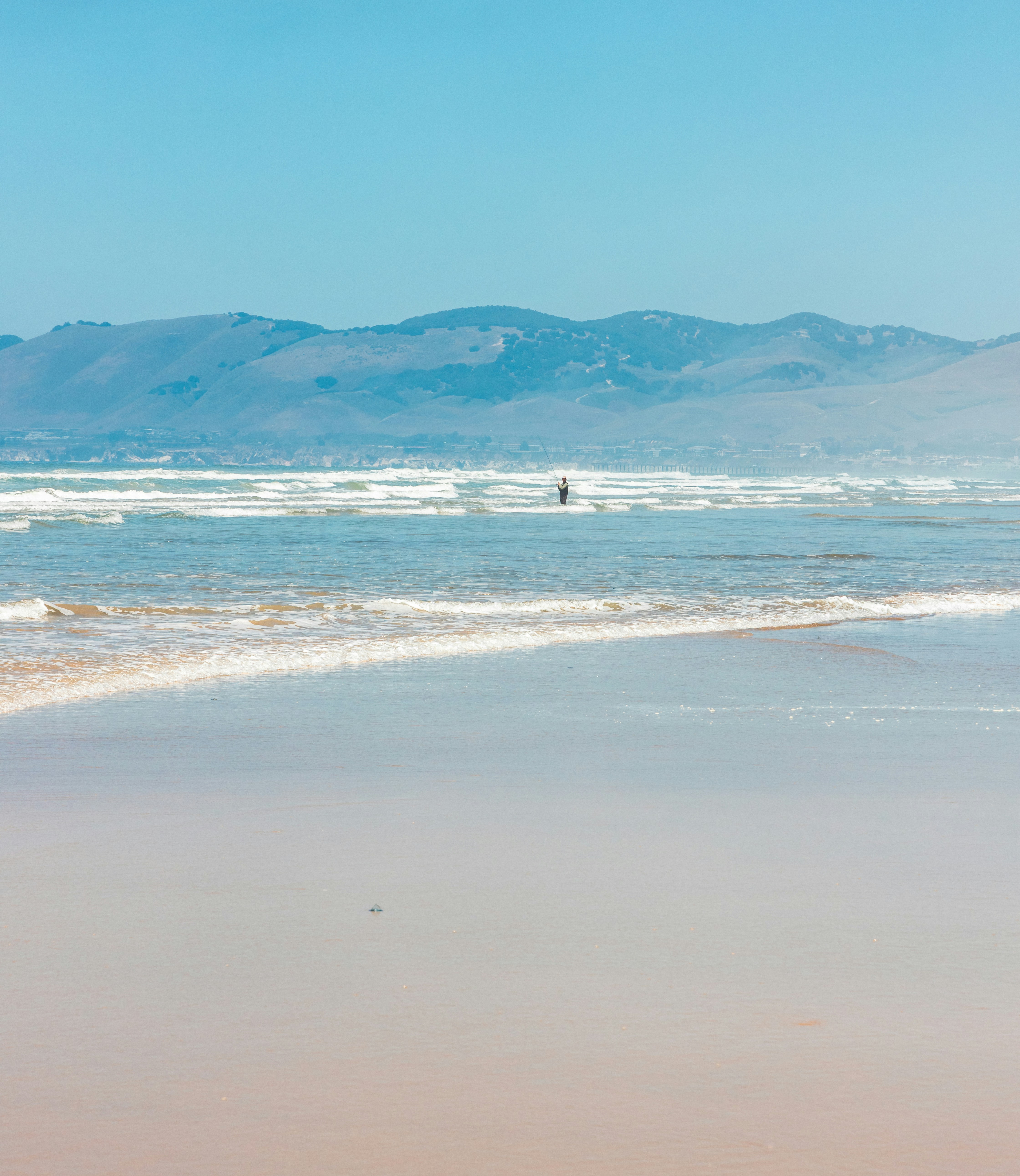 A person fishing in the surf before distant rolling hills.