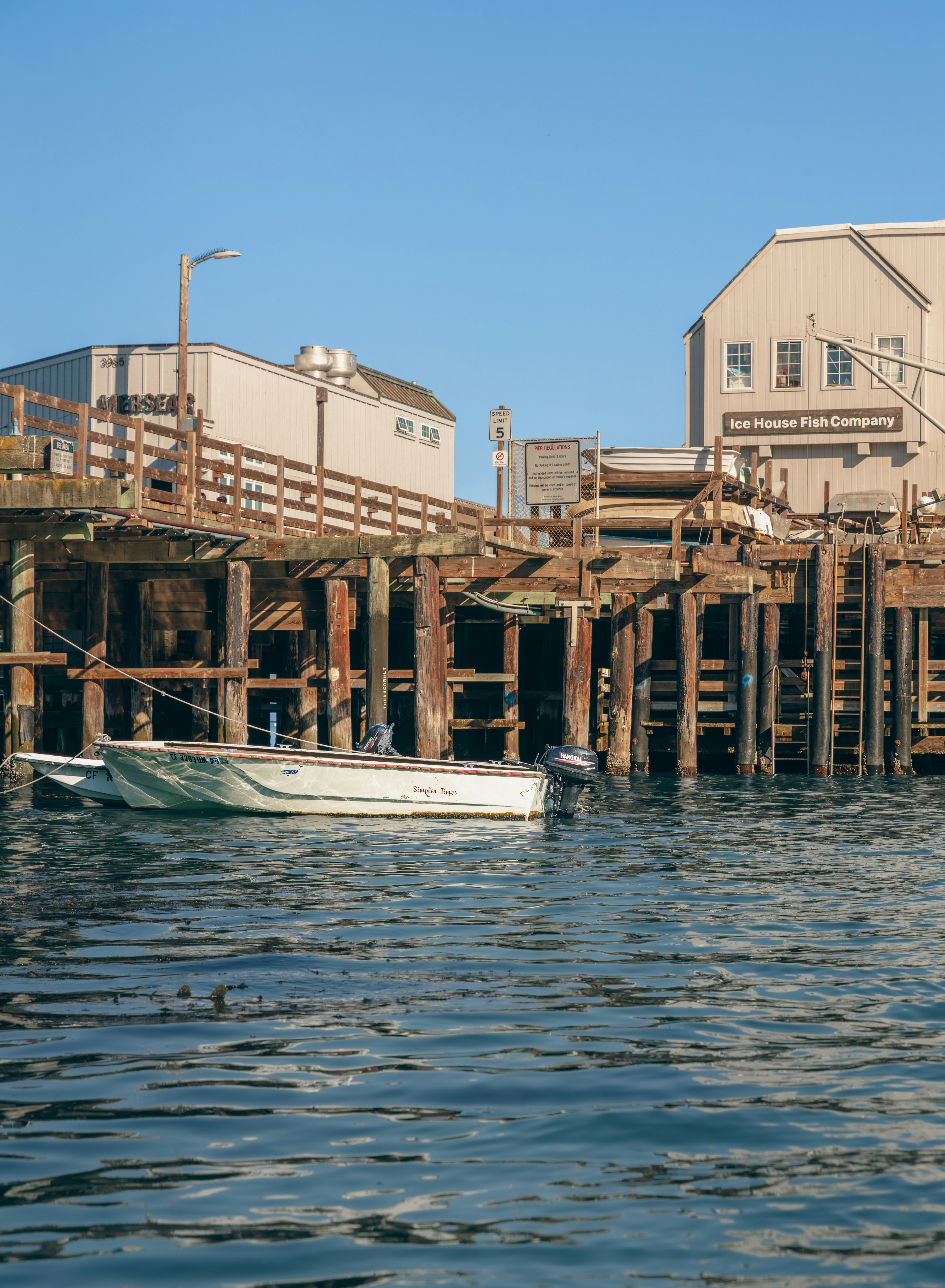 A small white motorboat moored at a wooden pier with buildings in the background.