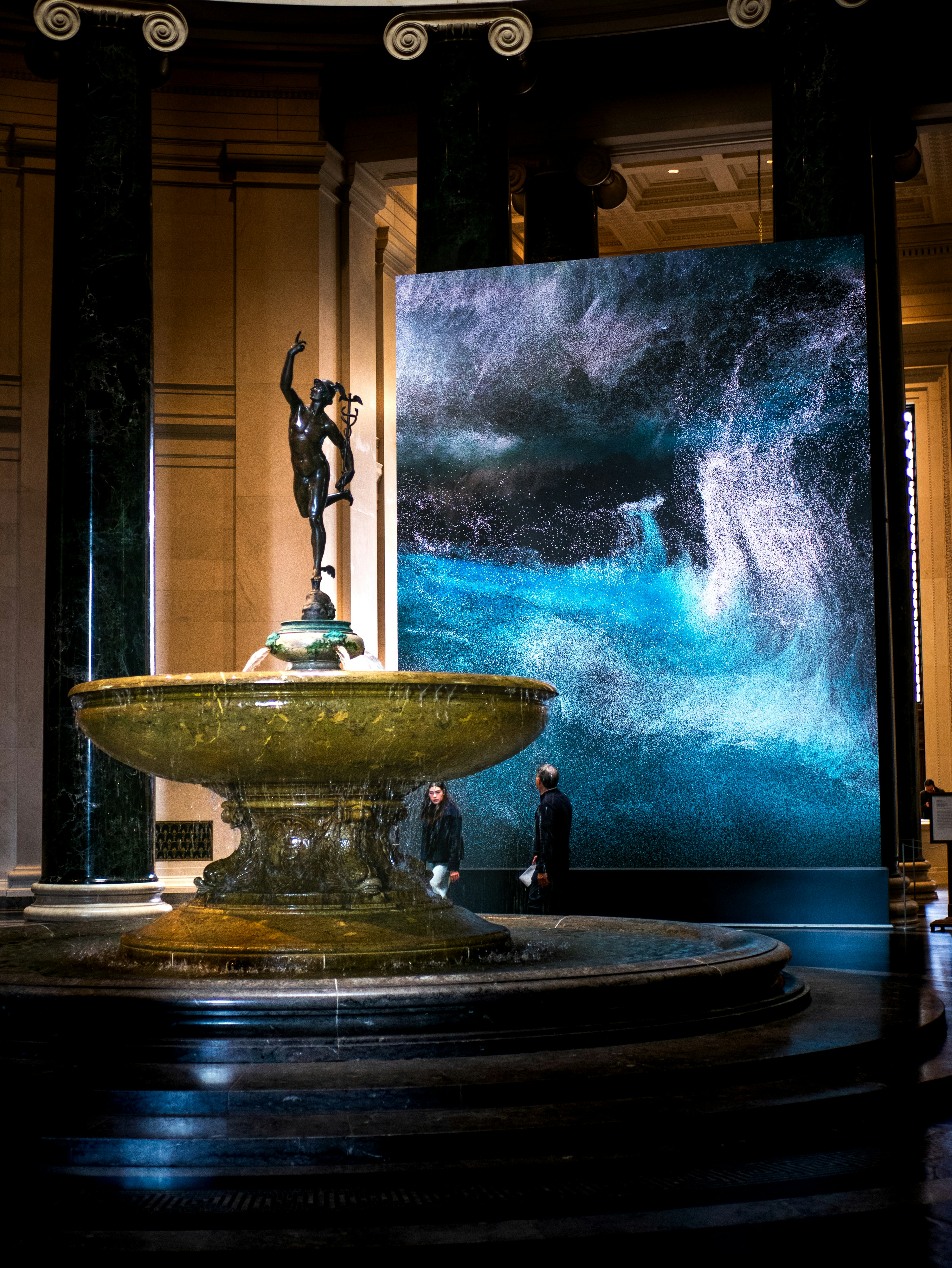 A bronze statue of Mercury above a fountain near a large digital ocean display