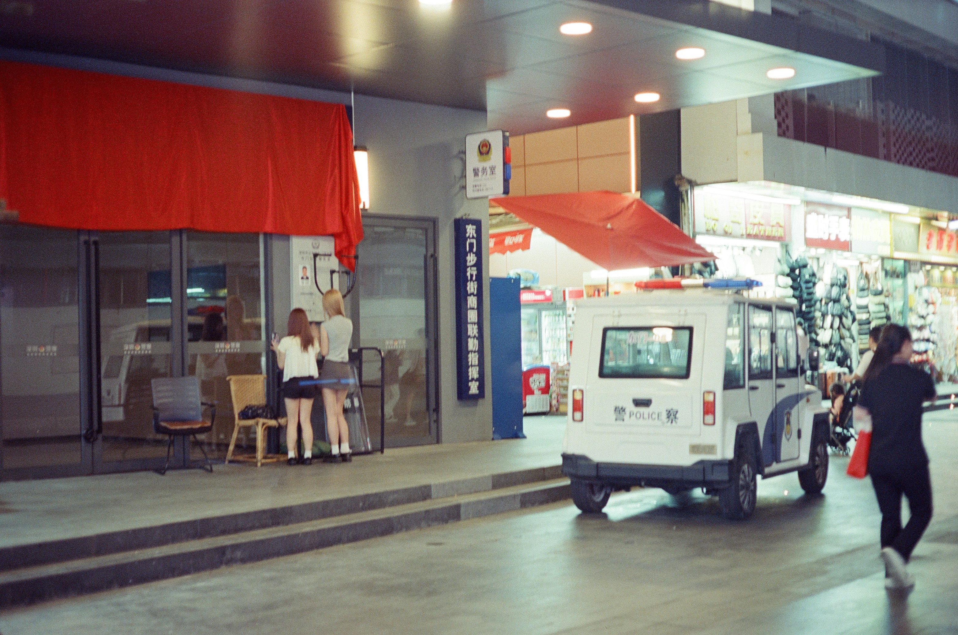 Two people standing before a police station entrance with a white patrol vehicle nearby.