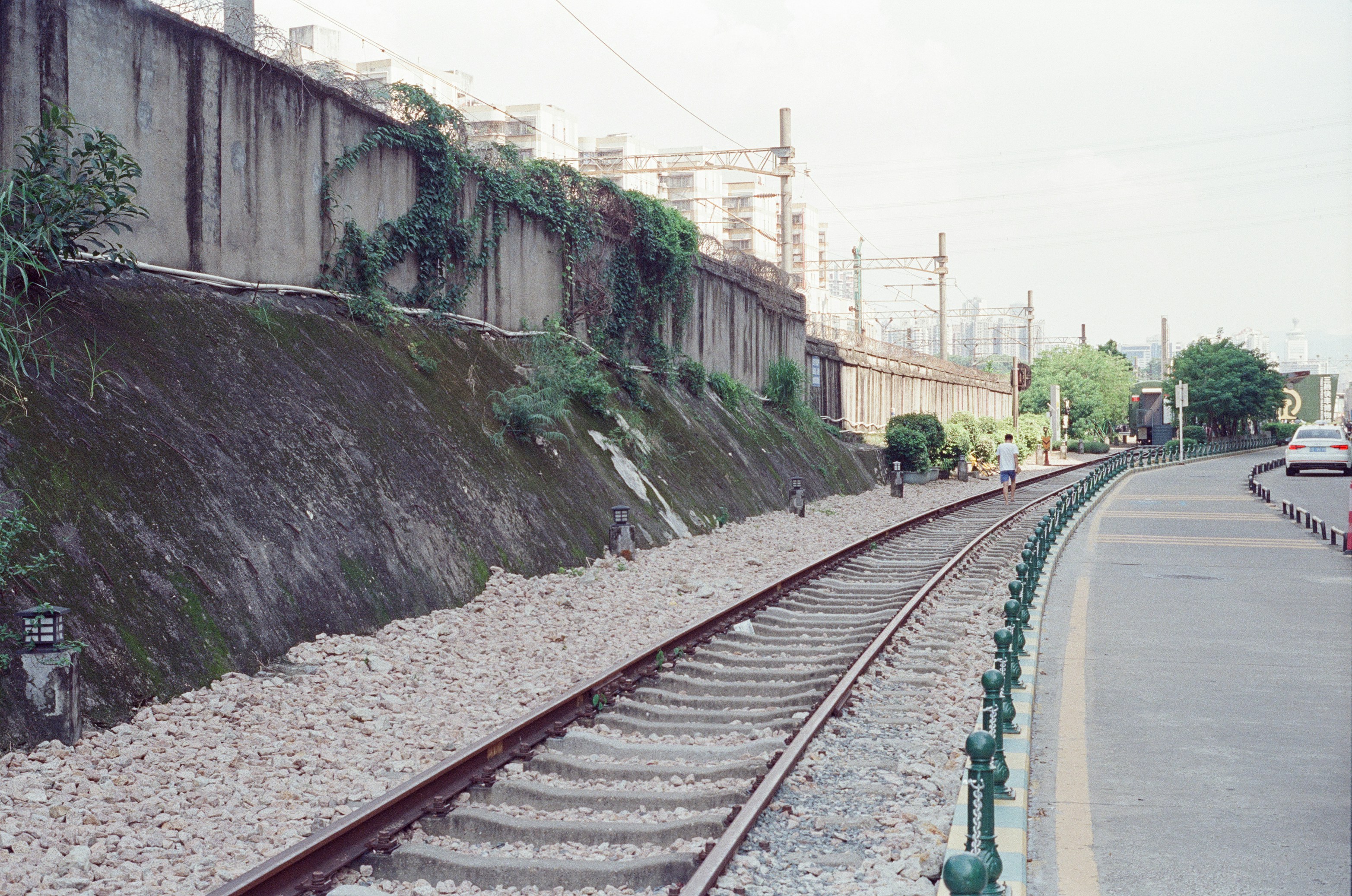 A person walking along railway tracks beside a concrete retaining wall with climbing vines.