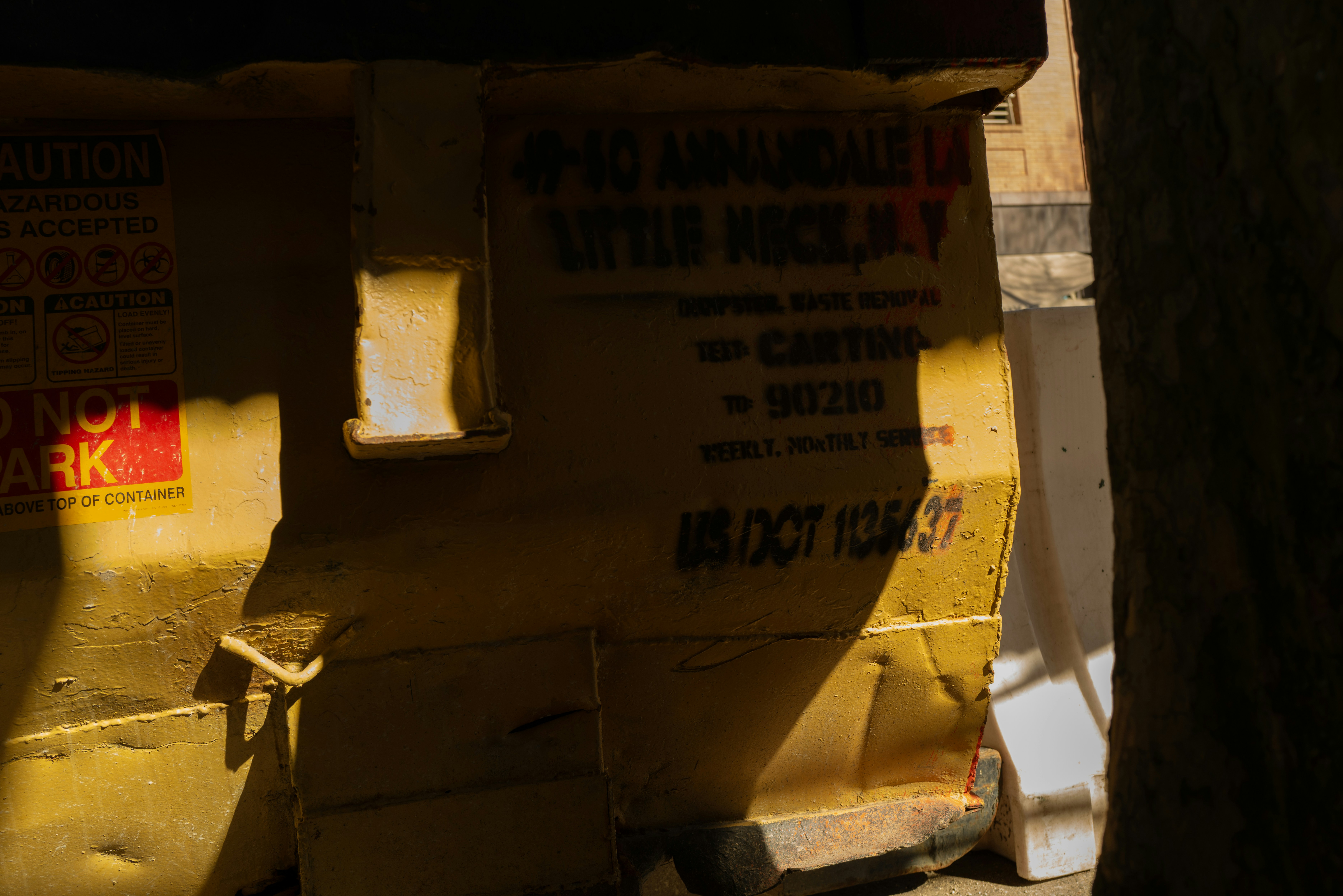 A yellow metal dumpster with stenciled text and warning labels in bright sunlight