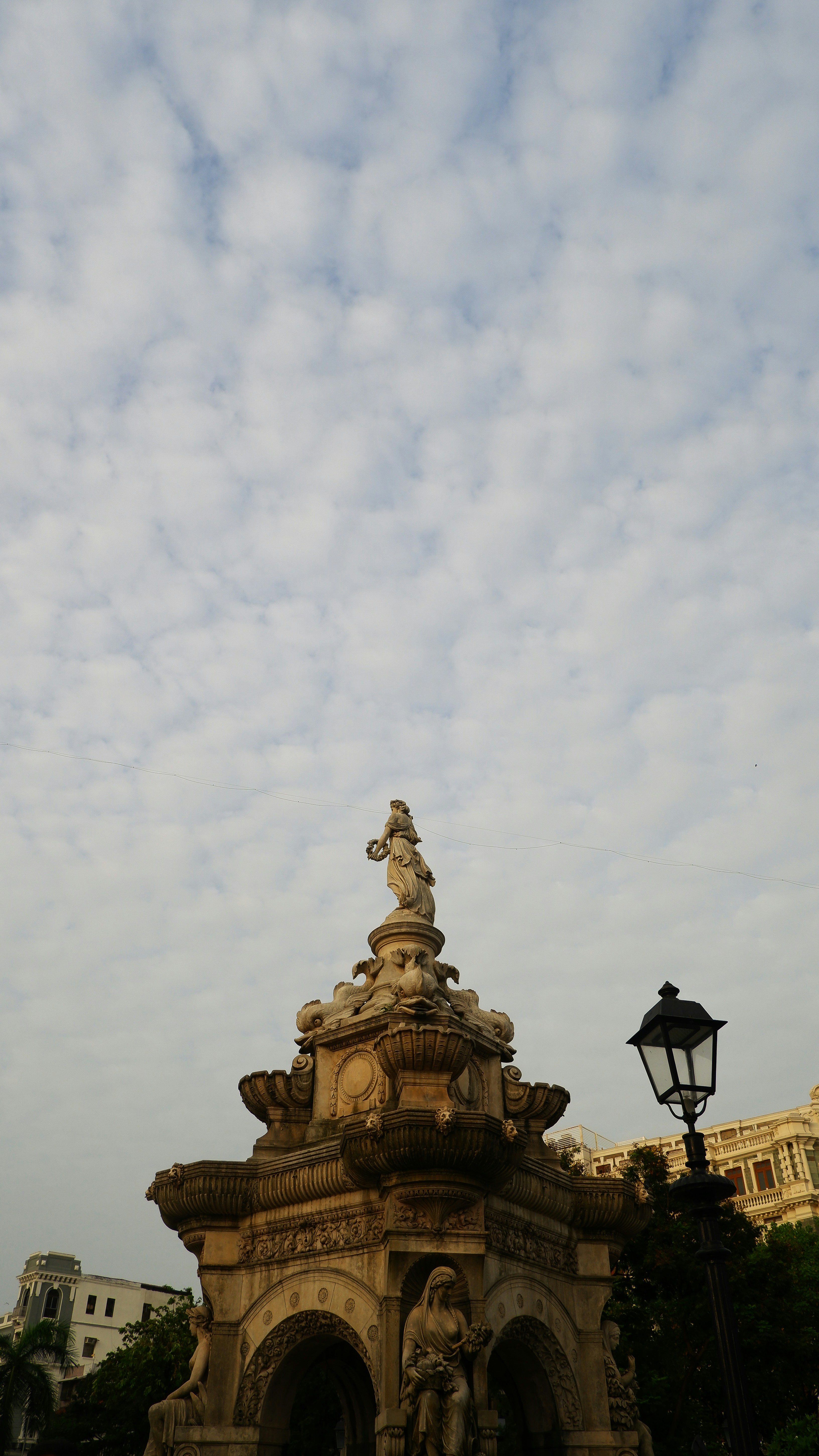 A stone fountain with classical statues under a sky filled with scattered clouds.
